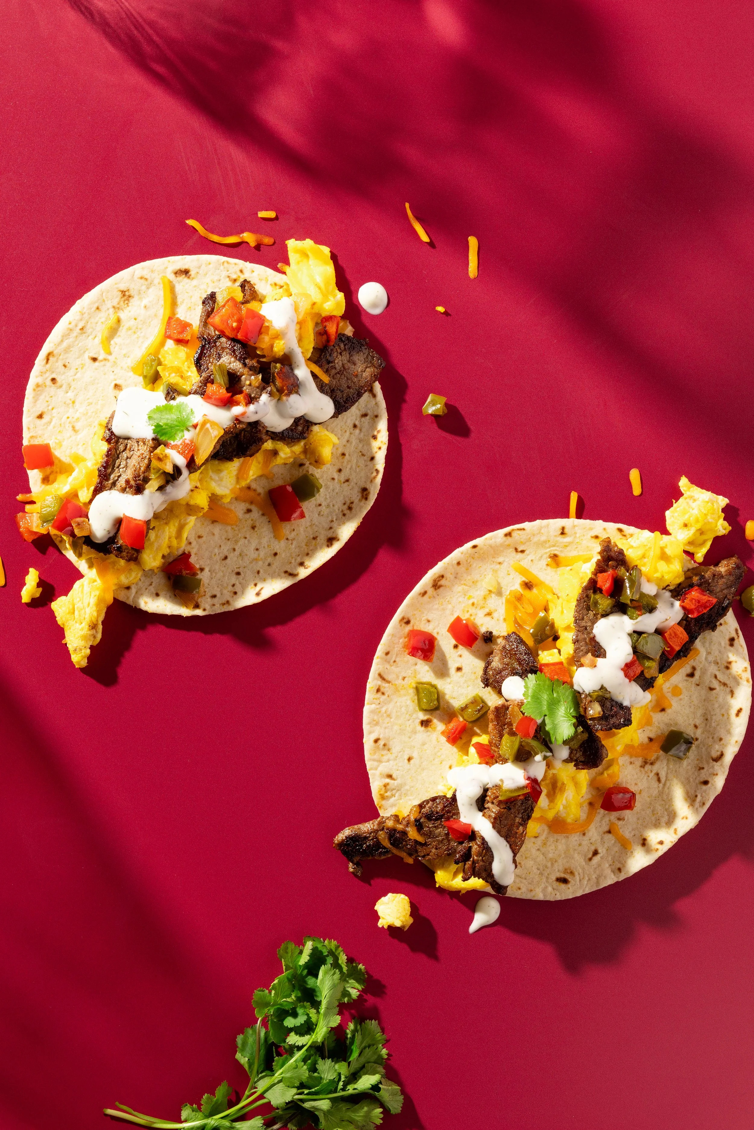 Two tacos with grilled meat, scrambled eggs, chopped vegetables, shredded cheese, cilantro, and creamy sauce on a red surface.