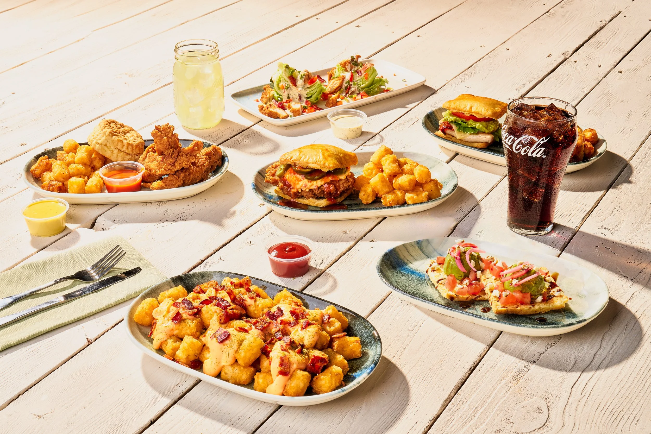 An assortment of fast food dishes on a white wooden table, including fried chicken with tater tots, a sandwich with lettuce and tomato, pizza slices with toppings, salad, and soft drinks.