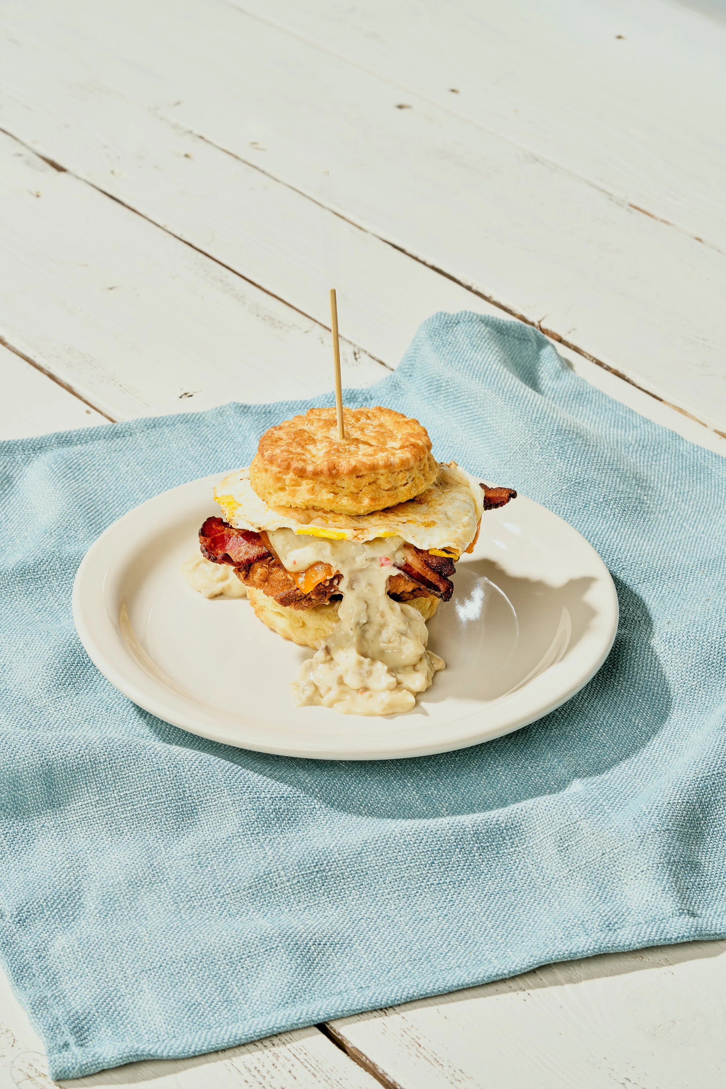 A plate with a breakfast sandwich made of a biscuit, fried egg, bacon, and gravy, topped with a biscuit and a toothpick, on a blue napkin on a white wooden surface.