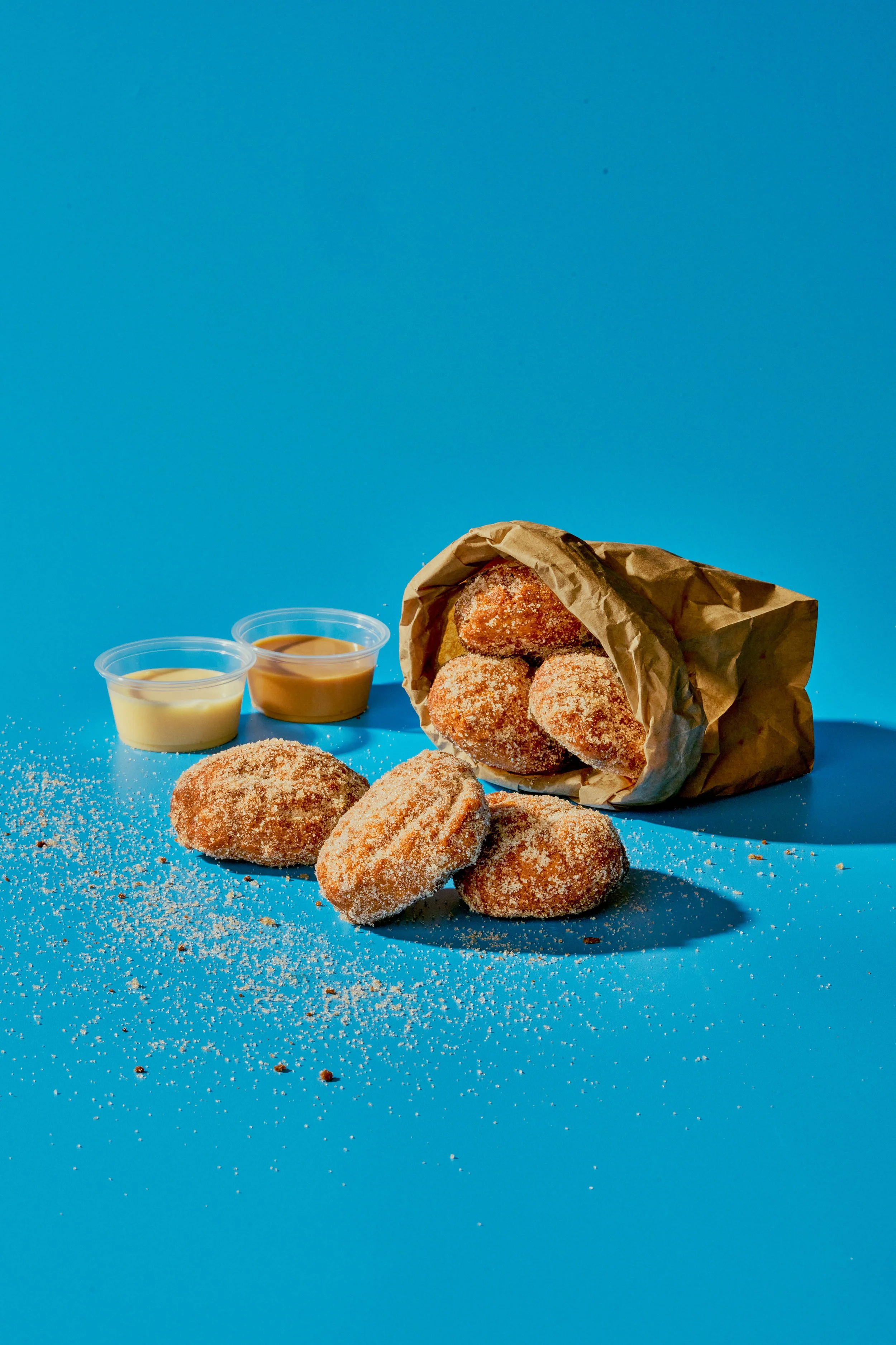 Crispy breaded chicken nuggets on a blue surface with two dipping sauces and a paper bag of more nuggets, some spilled onto the surface.