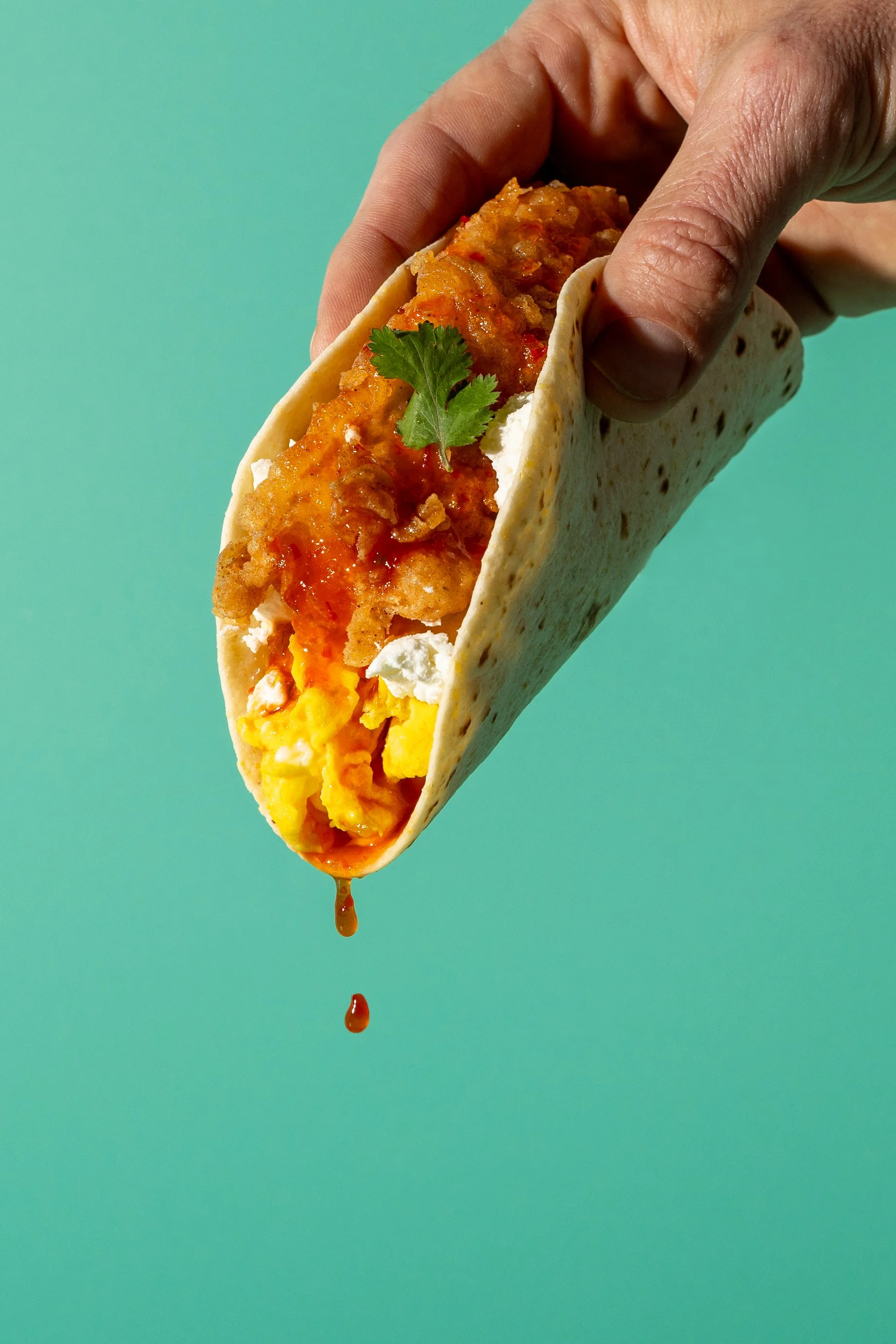 Hand holding a breakfast taco filled with scrambled eggs, fried chicken, hot sauce, and garnished with cilantro, with hot sauce dripping from the taco.
