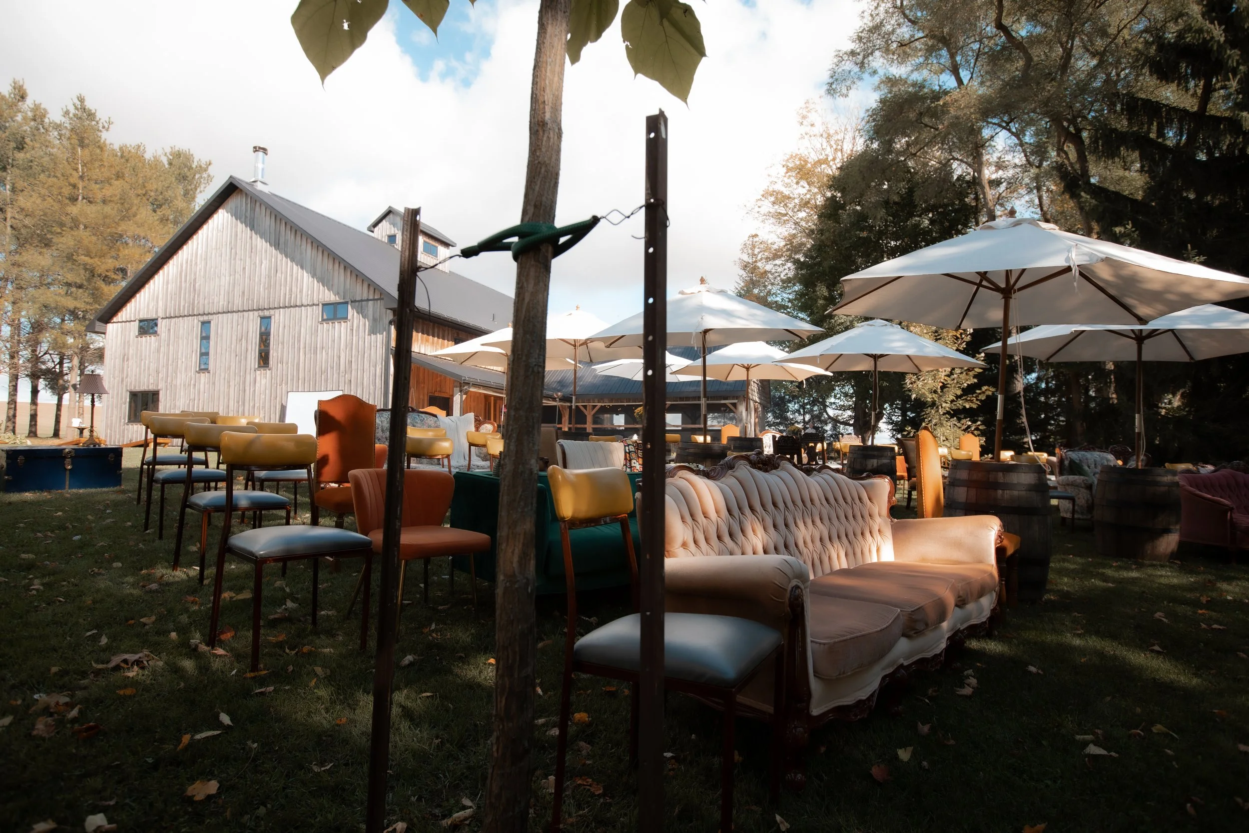 Outdoor seating area with vintage sofas, chairs, and white umbrellas in front of a rustic wooden building surrounded by trees.