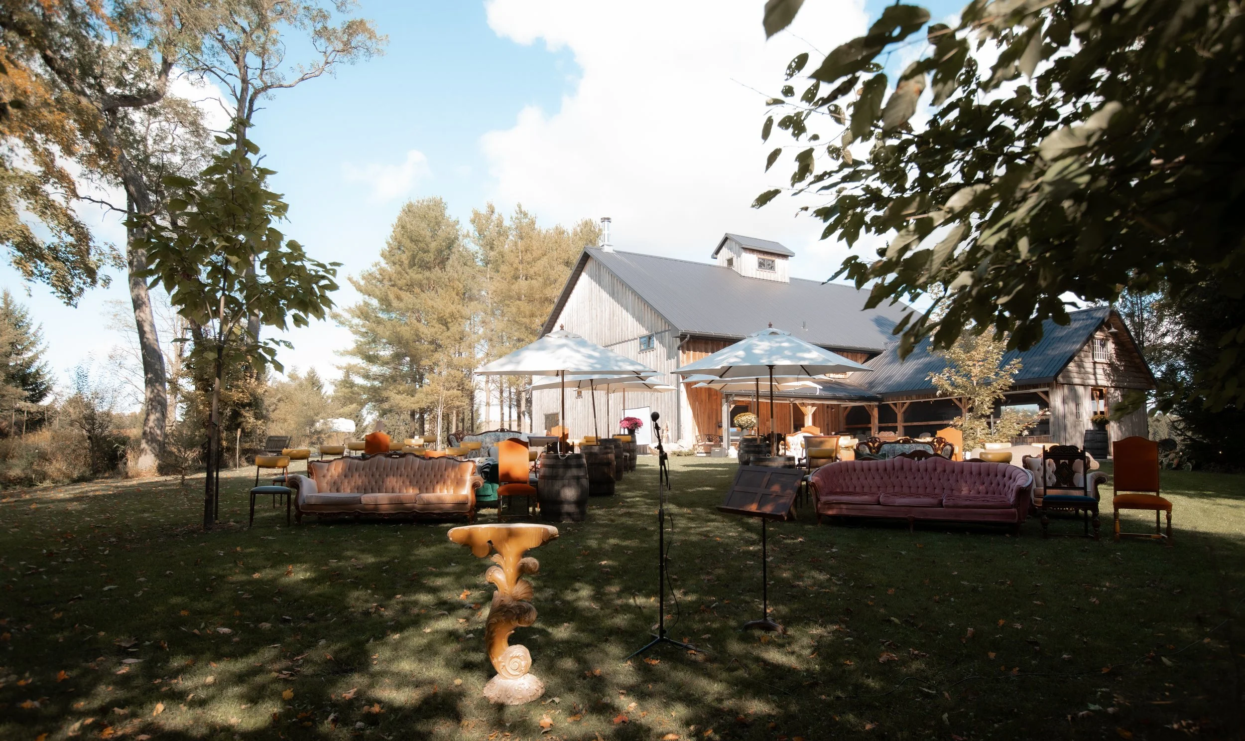 Outdoor setup with vintage sofas, chairs, umbrellas, and music stands on a lawn in front of a rustic barn during daytime.