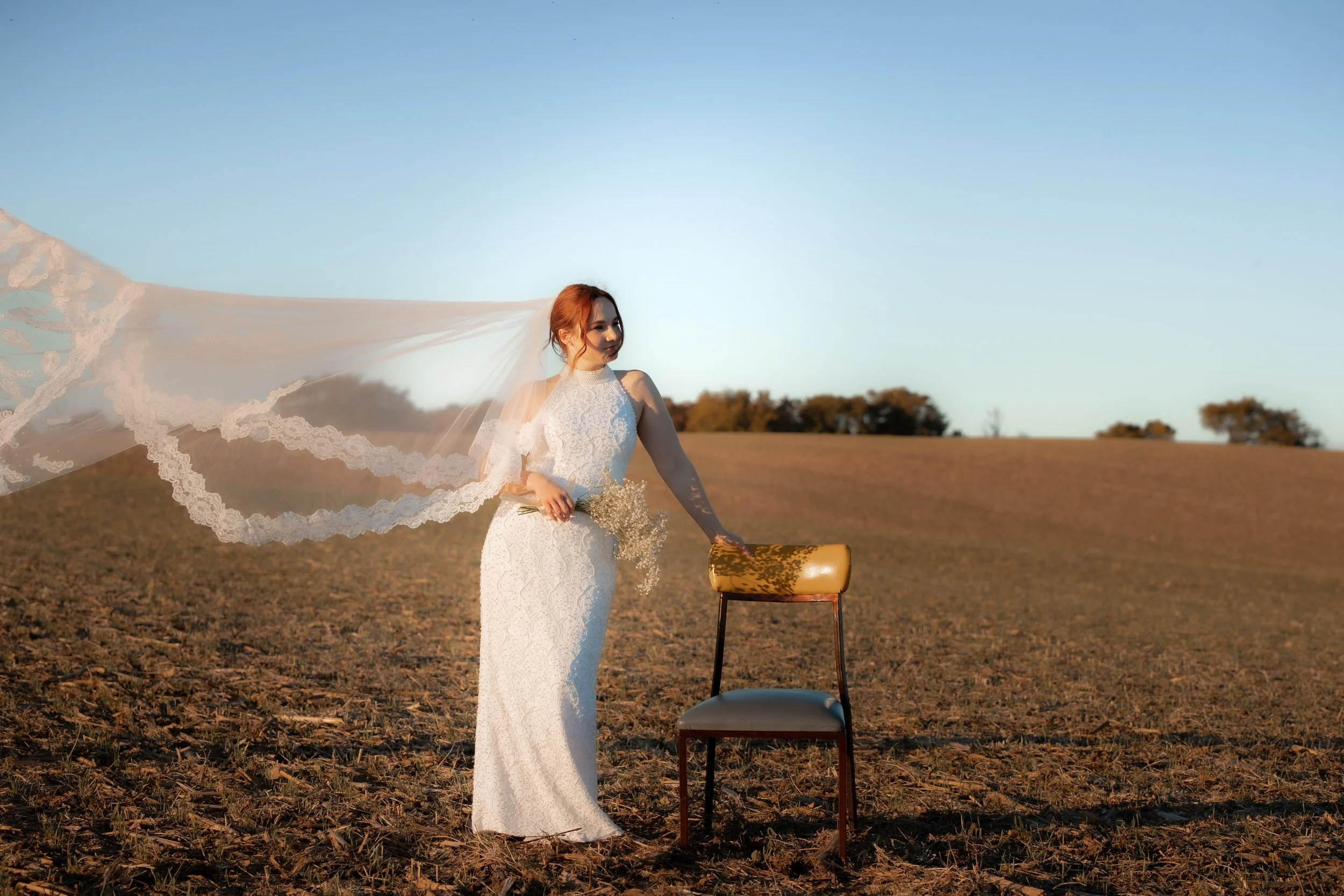 A woman in a white lace wedding dress and veil standing outdoors in a field, with her hand resting on the back of a yellow upholstered chair, under a clear blue sky at sunset.