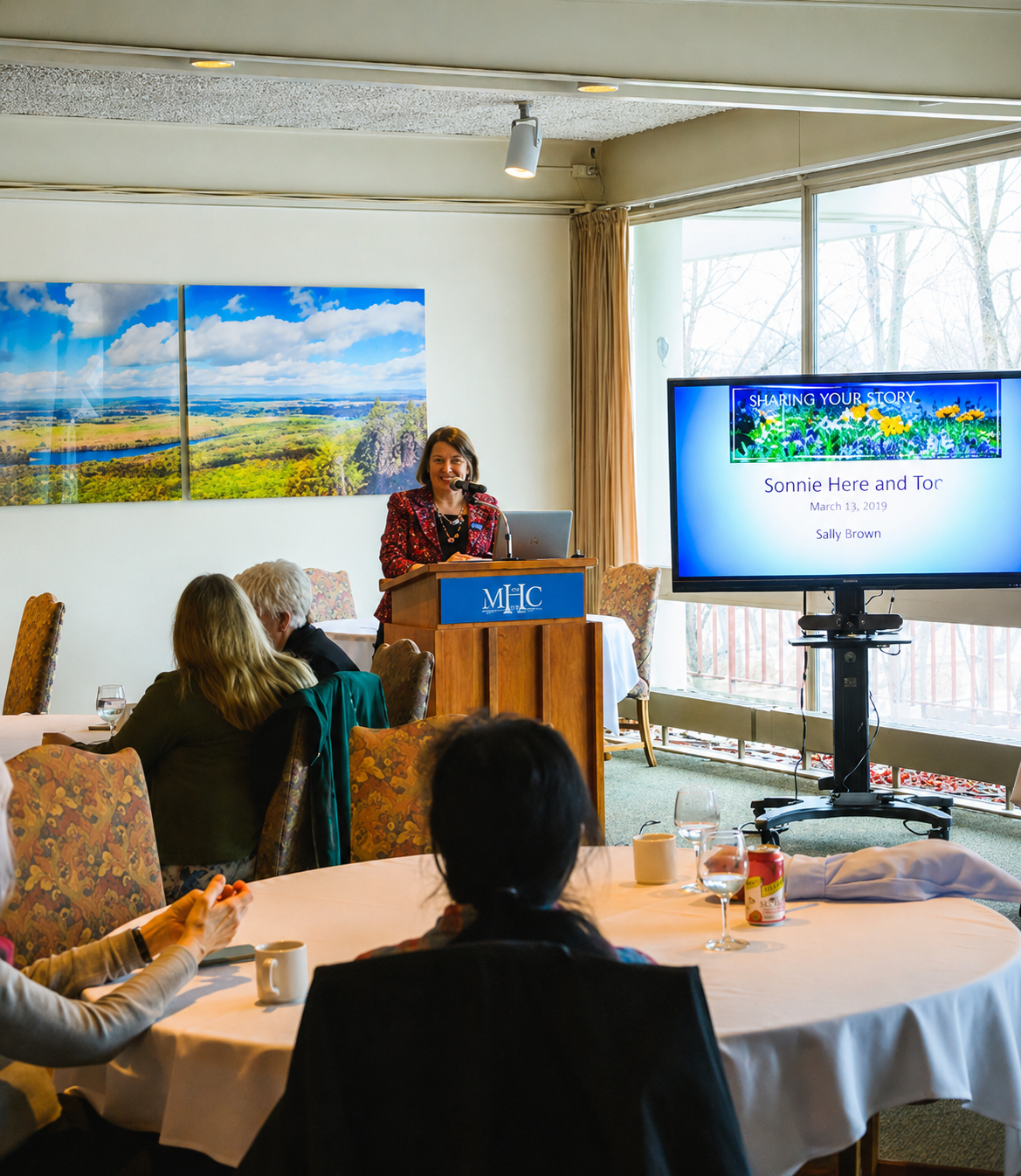 A woman giving a presentation at a podium with a microphone, in a room with round tables, each set with glasses and cups. A large screen displays a slide titled "Sharing Your Story" with the name Sonnie Here and Tor, dated March 13, 2019, by Sally Brown. Several people are seated, listening to the speaker. There is a panoramic landscape painting on the wall behind her and large windows with beige curtains showing trees outside.