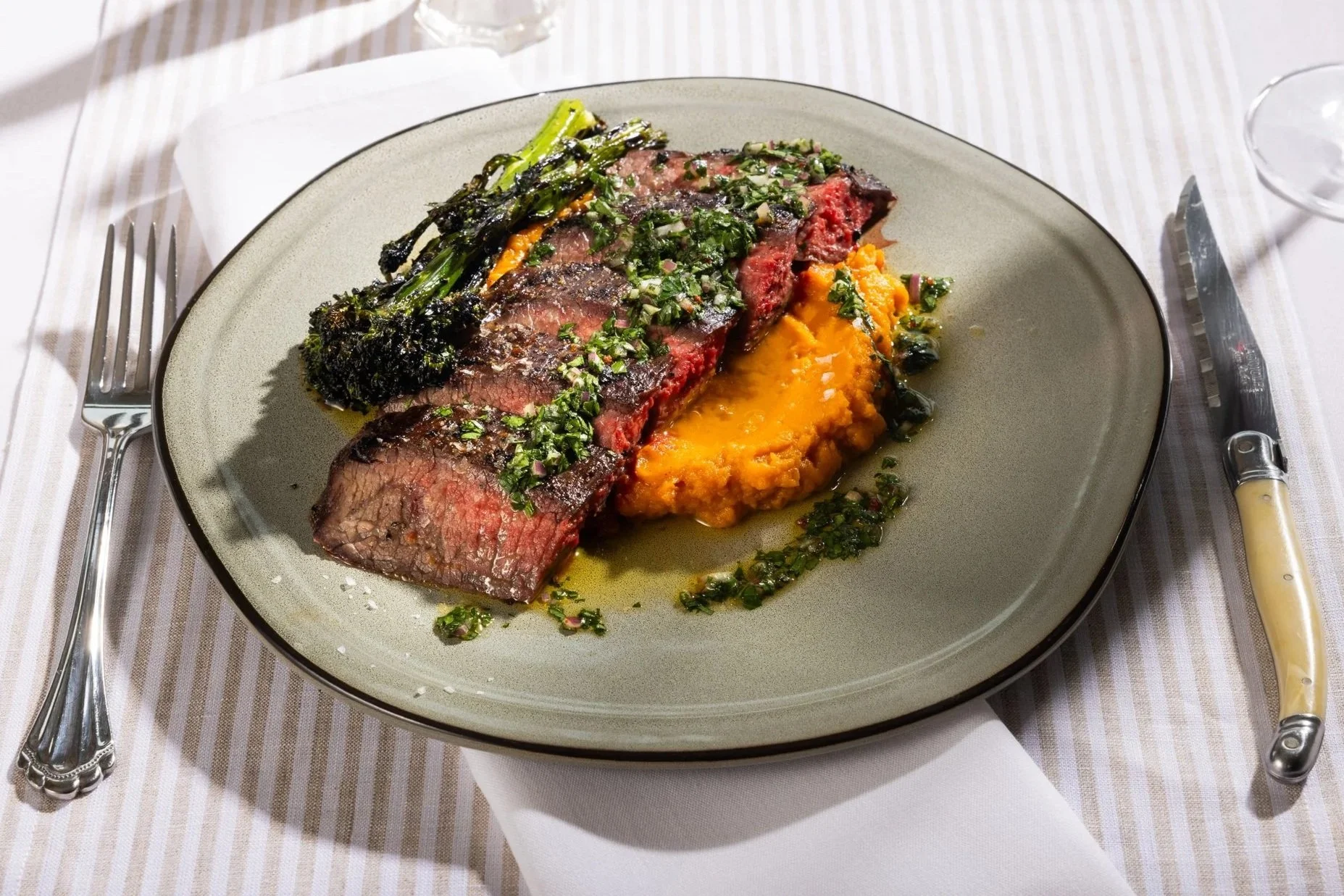 A plate of medium-rare beef steak topped with chopped herbs, served with roasted broccolini and mashed sweet potatoes.