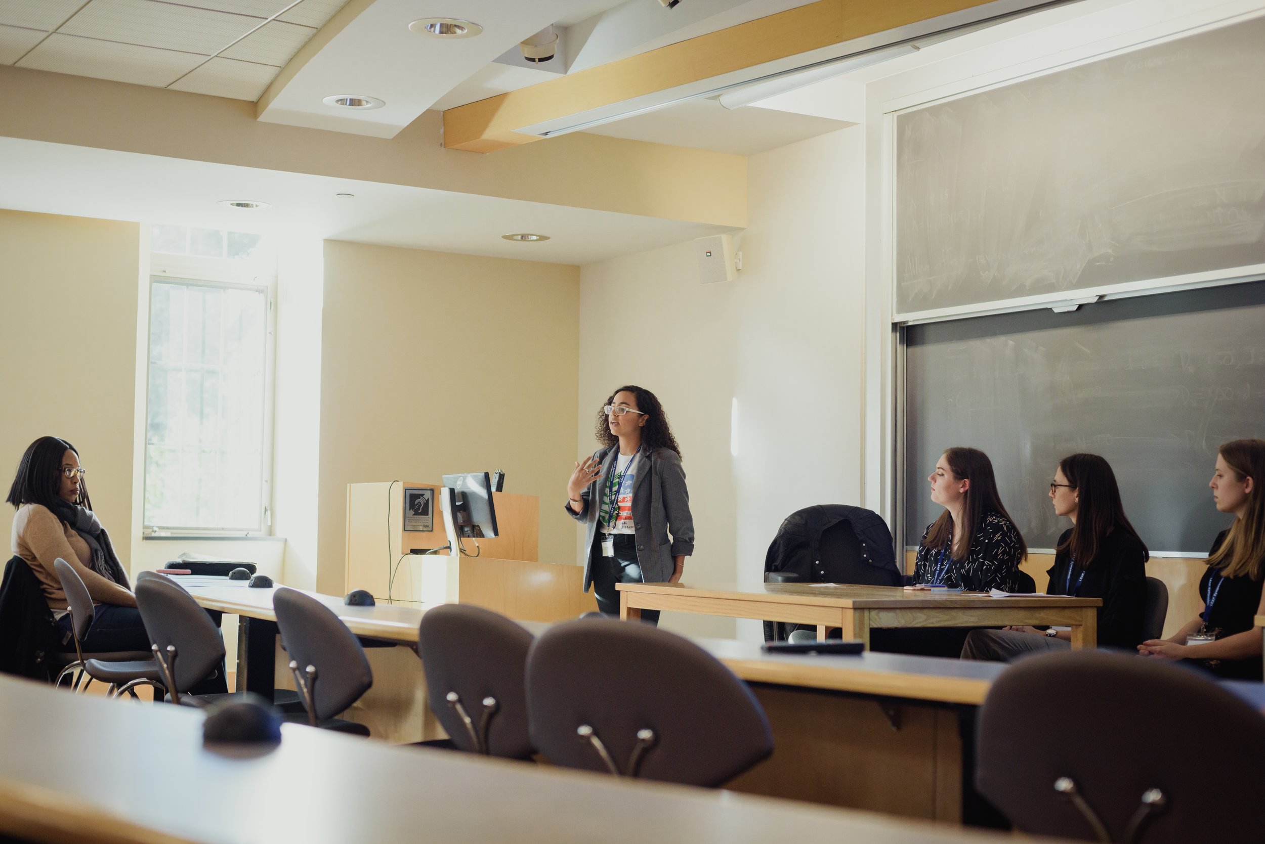 A woman standing and speaking to a group of four women sitting at a conference table in a classroom or lecture hall.