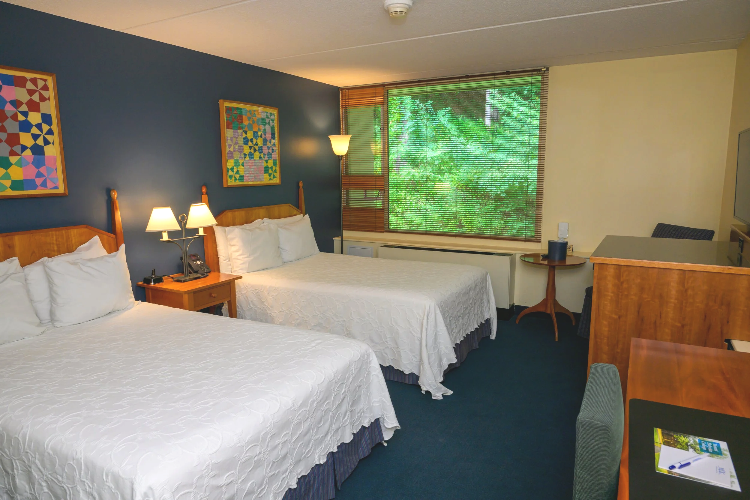 Hotel room with two beds, wooden headboards, nightstand with lamps and phone, large window with blinds and greenery outside, small round table, TV on a wooden dresser, and a desk with a chair.