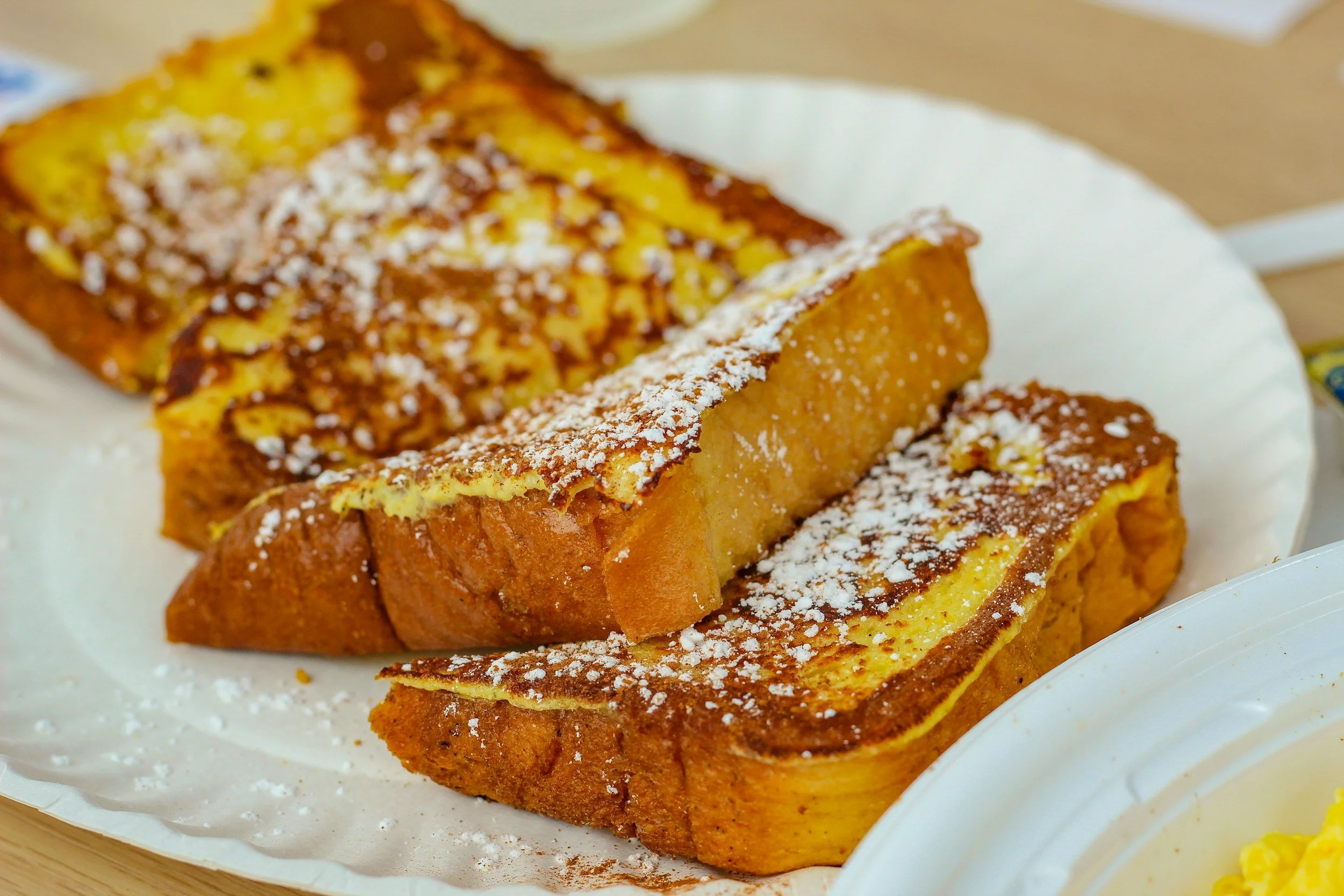 Close-up of French toast covered with powdered sugar on a white plate, with a side of scrambled eggs in a small dish.