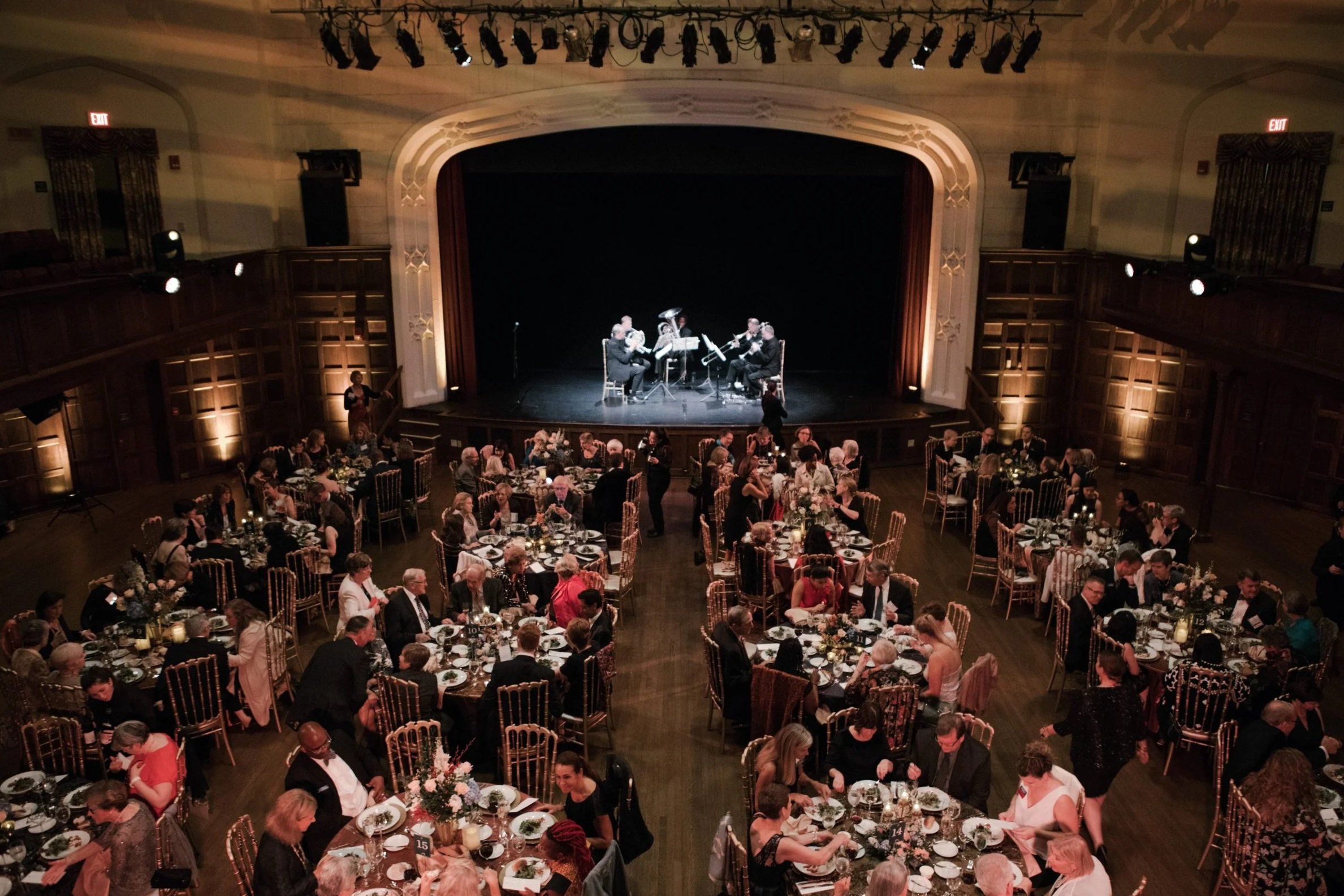 A large banquet hall filled with round tables and people dining, with a stage at the front where a small musical ensemble is performing.