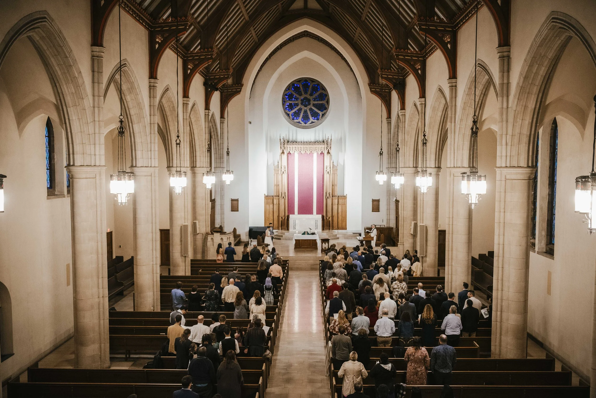 People gathered in a church interior for a service or ceremony, with high vaulted ceilings, stained glass windows, and chandeliers.