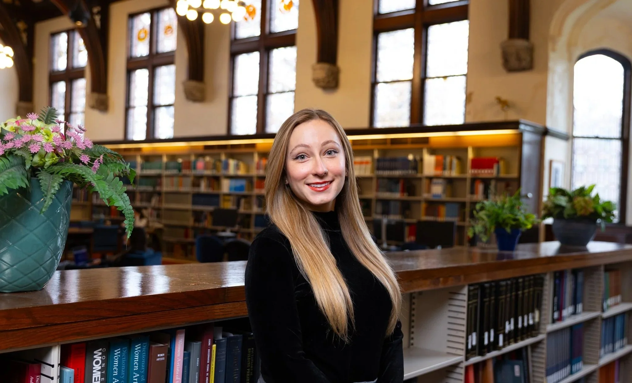 A young woman with long blonde hair smiling in a library, with large windows and shelves full of books in the background.