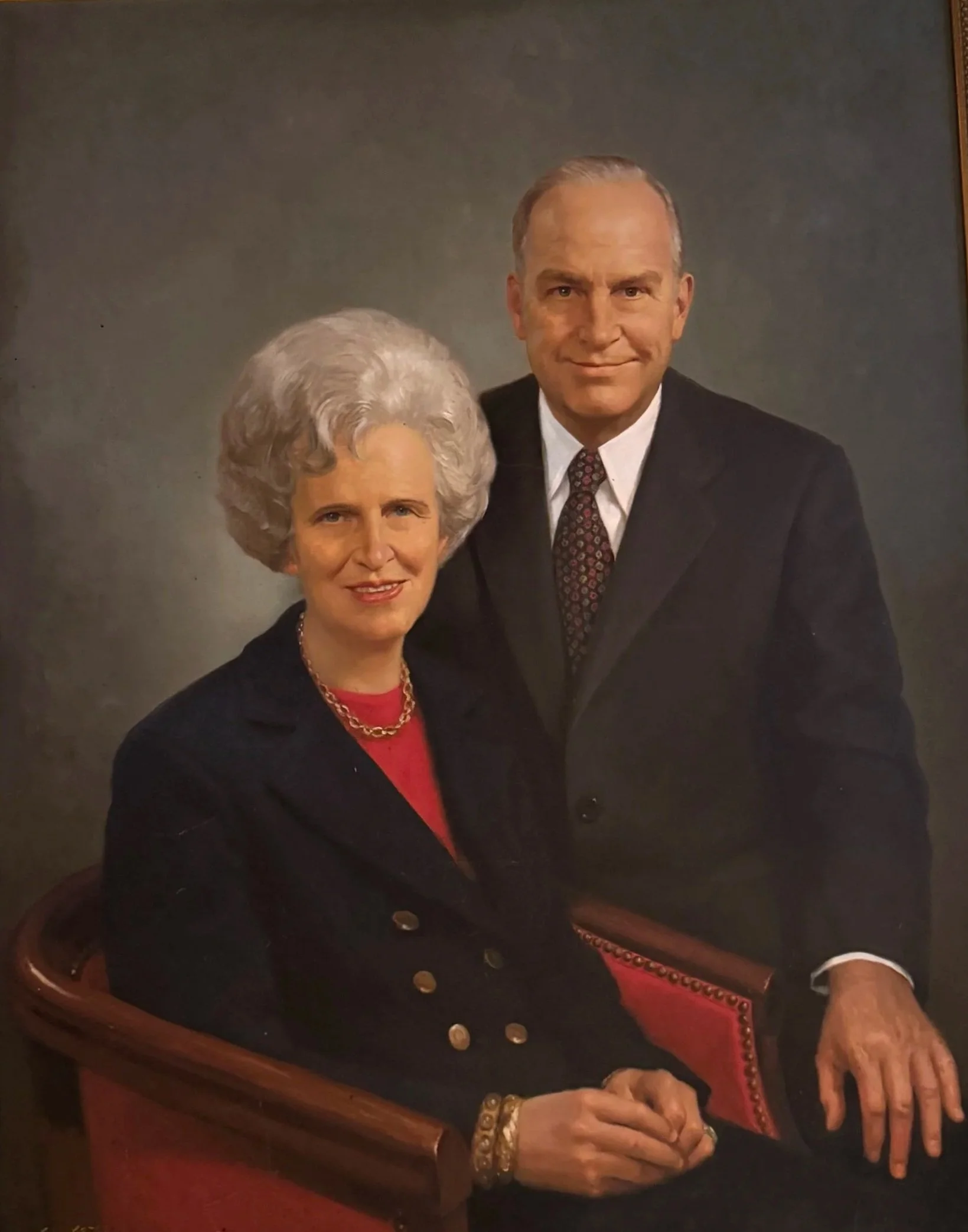 A portrait of an elderly woman with styled white hair, wearing a black blazer over a red top, gold jewelry, and sitting at a wooden table. Adjacent to her, a man with blond hair, dressed in a dark suit, white shirt, and patterned tie, is standing and smiling.