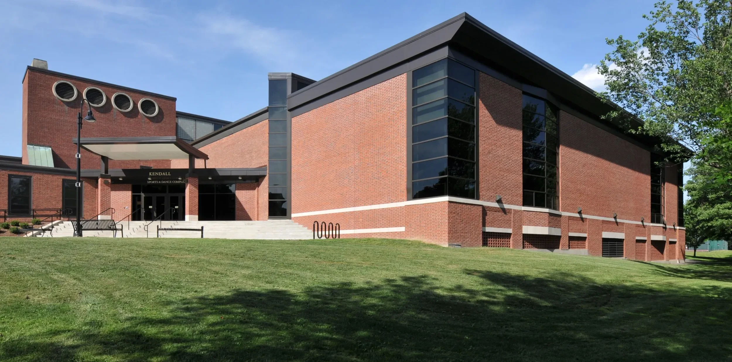 A brick building with large windows, stairs leading to the entrance, and a sign reading 'Kendall Sports & Dance Complex.' The building is surrounded by green grass, trees, and a clear blue sky.