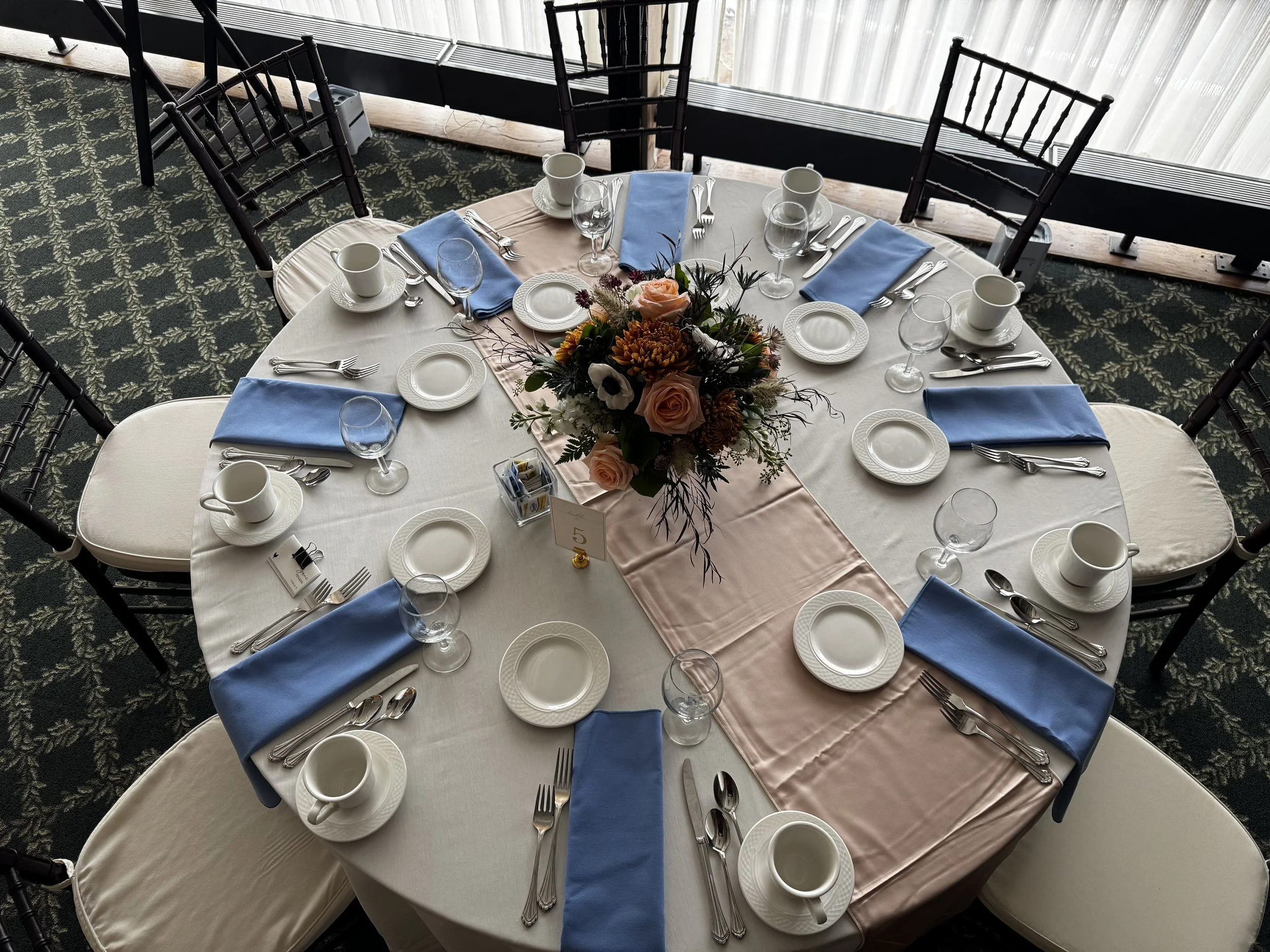 Round banquet table set with white tablecloth, blue napkins, white plates, silverware, wine glasses, coffee cups, and a floral centerpiece with pink roses and other flowers, in a room with patterned carpet and large windows with sheer curtains.
