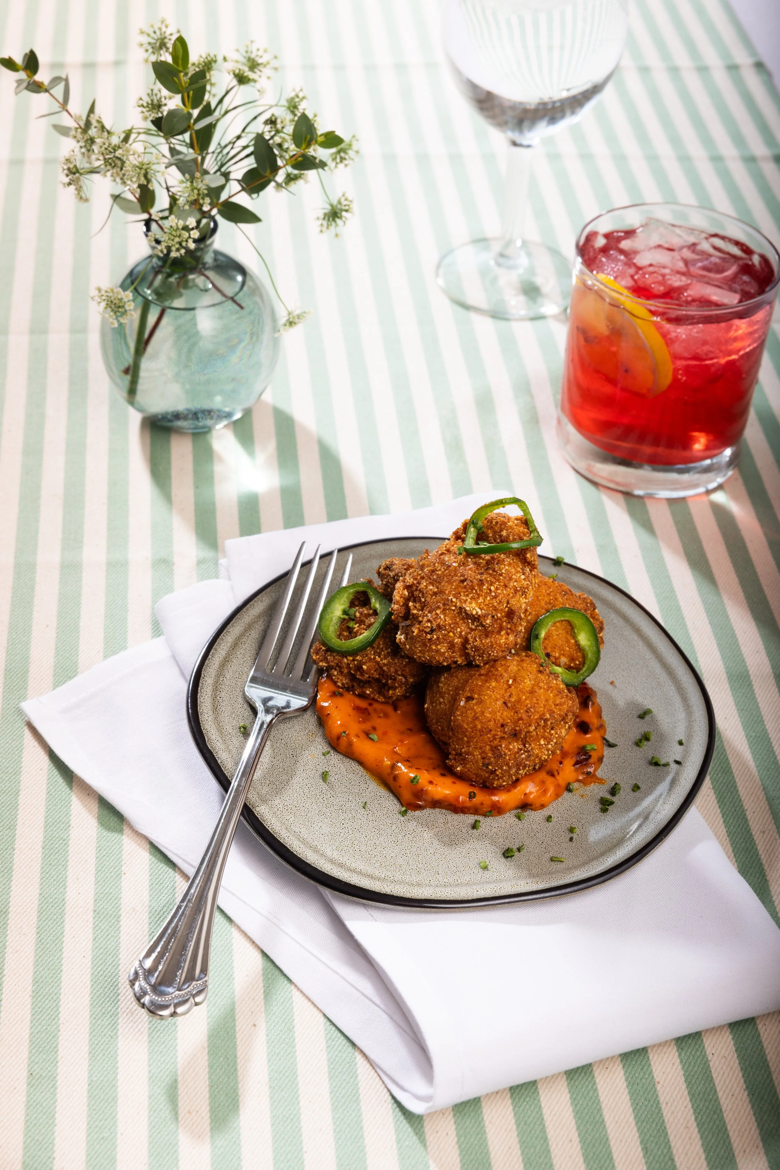 A plate of fried chicken drizzled with orange sauce, garnished with sliced green peppers, on a striped green and white tablecloth. In the background, a glass of red beverage with lemon slice, a glass of water, and a small vase with green leafy and wh