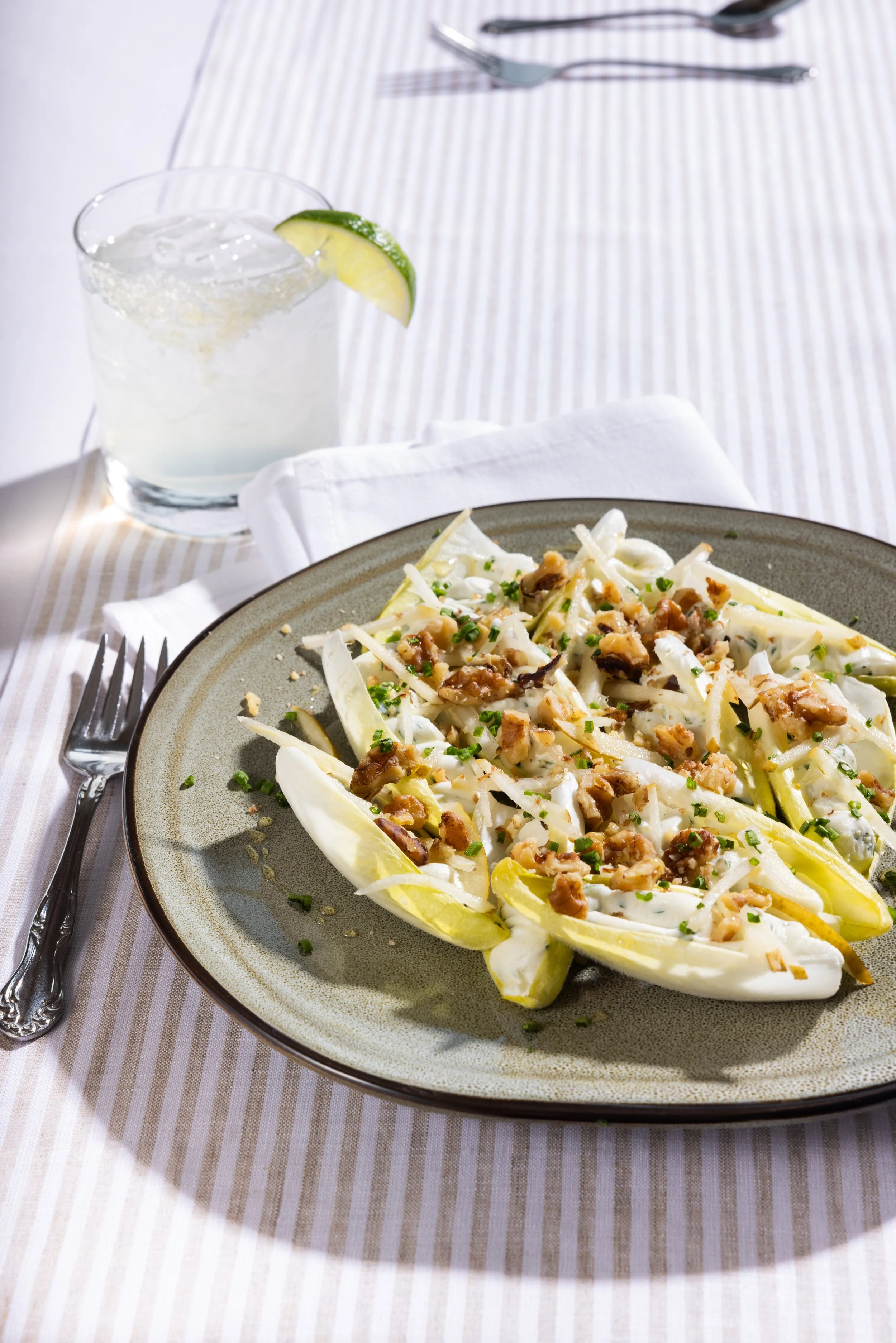 Halved endive lettuce filled with chopped nuts and chopped herbs, served on a round gray plate. In the background, a glass of sparkling water with ice and a lime wedge, set on a white and beige striped tablecloth, with silver utensils nearby.