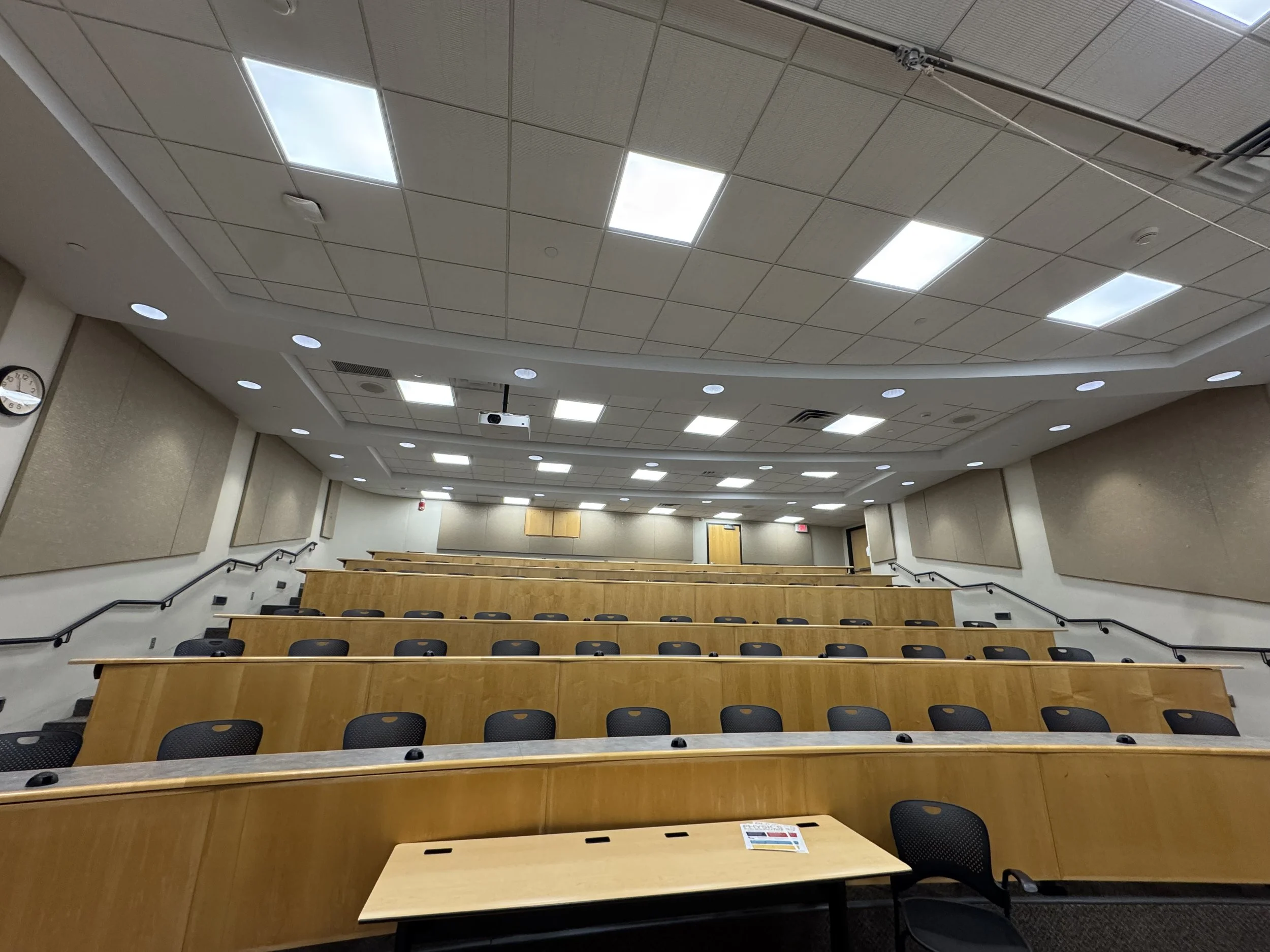 Empty lecture hall or classroom with tiered wooden desks and black chairs, beige walls, a clock, and a ceiling with bright overhead lights.