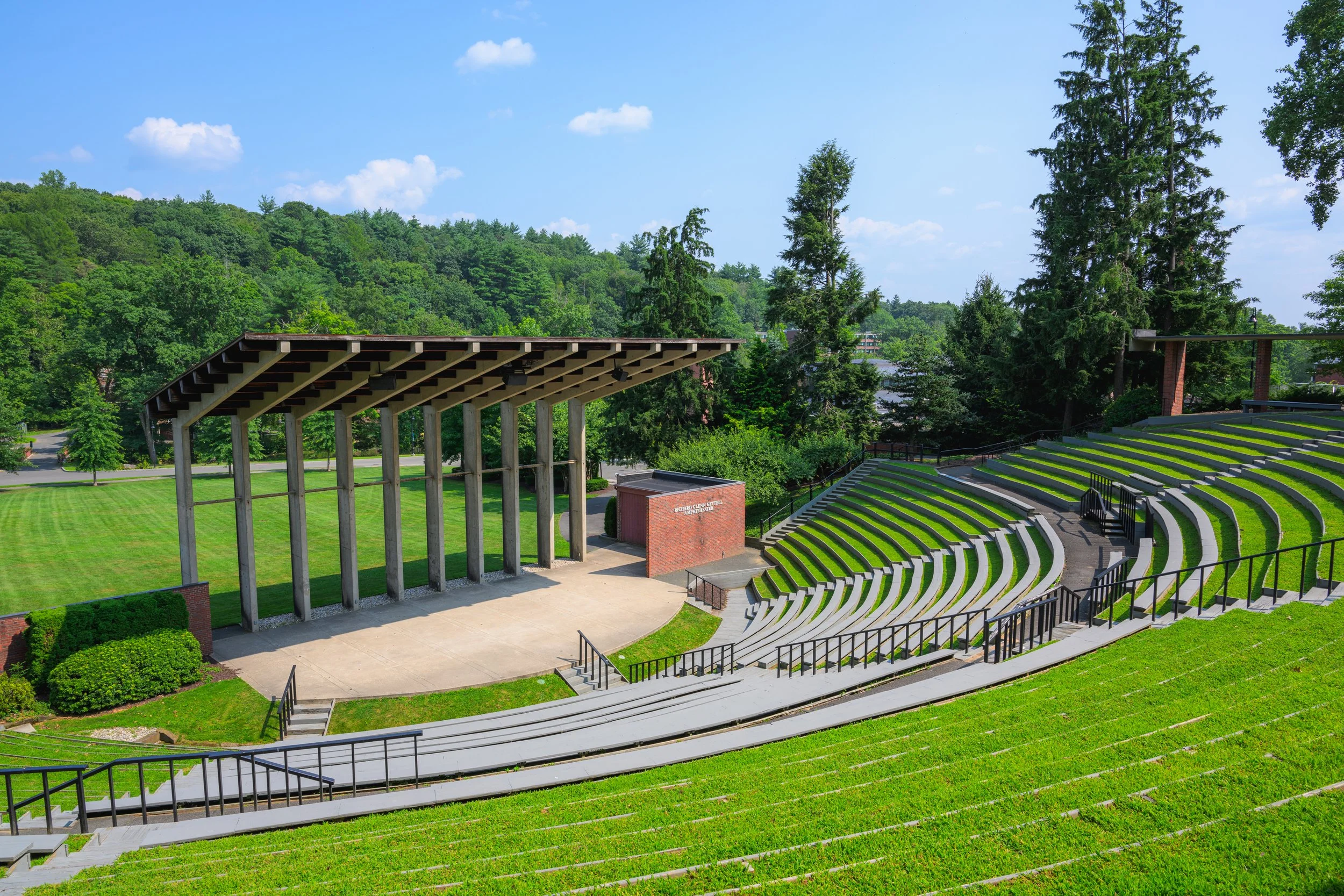 Outdoor amphitheater with tiered seating, a stage with a modern shade structure, surrounded by green trees and rolling hills under a blue sky.