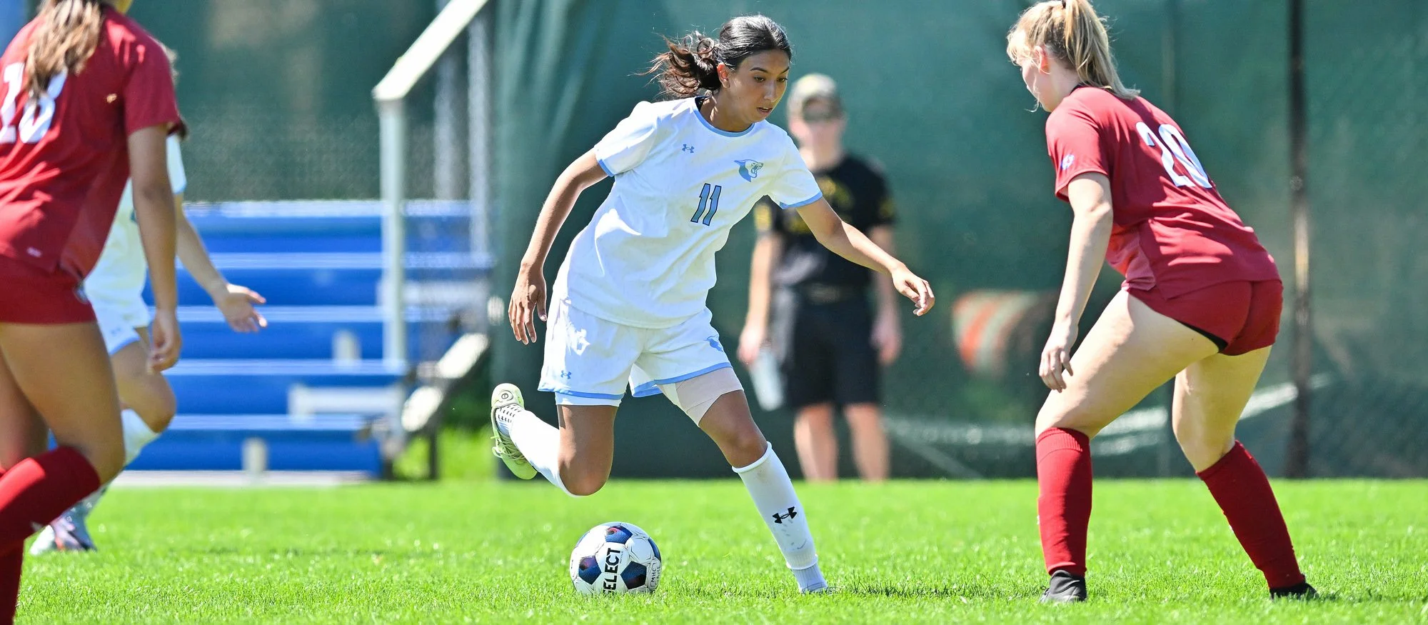 A female soccer player in a white uniform with the number 11 on her jersey dribbling a soccer ball while being defended by a female player in a red uniform with the number 20 on her shorts on a grassy field.