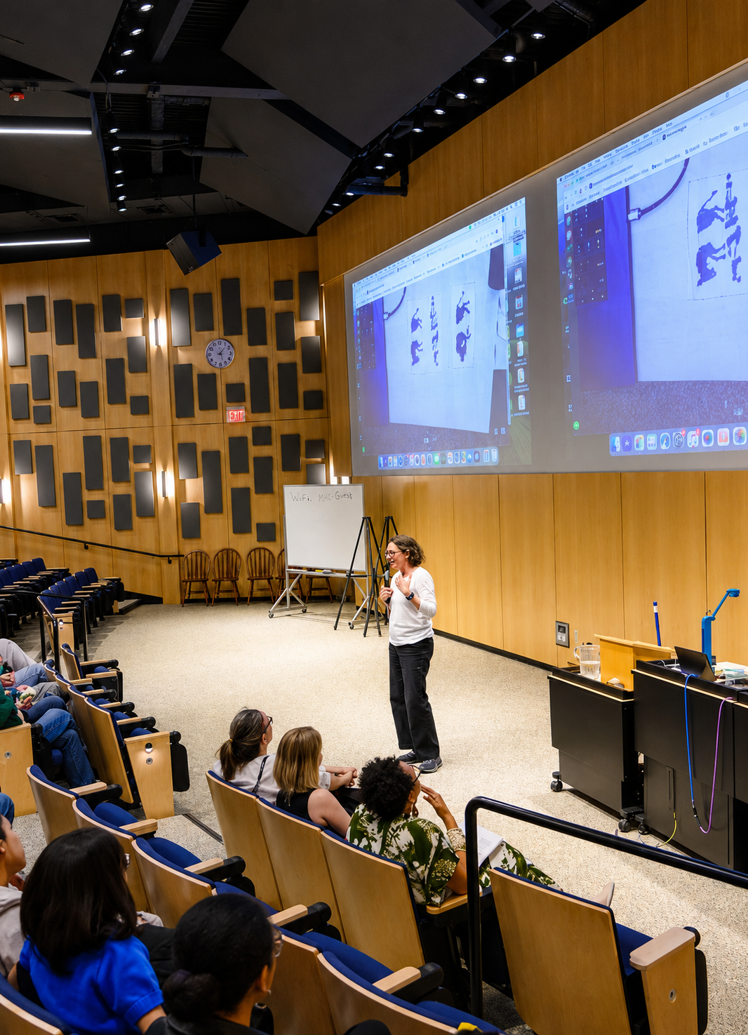 A woman giving a presentation in a lecture hall with a large screen displaying her slides, and a small group of people seated and listening.