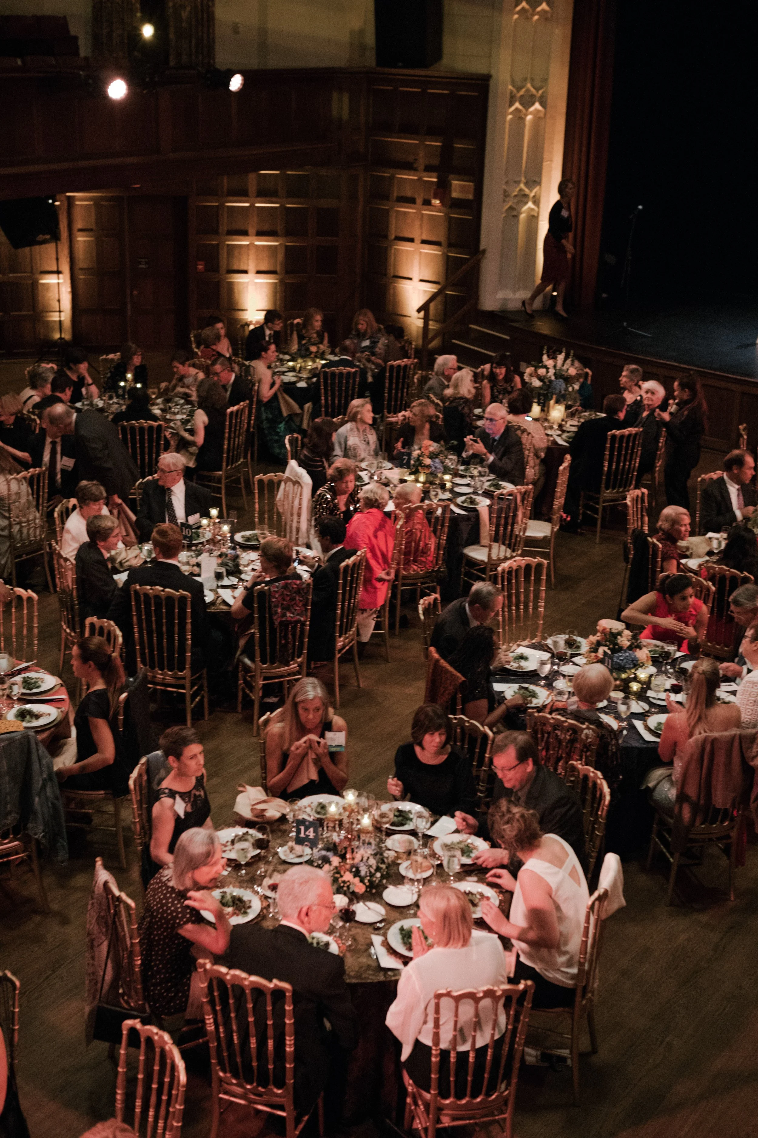 People seated at round tables with floral centerpieces, eating and conversing in a formal dining event with wood-paneled walls and stage with speaker.