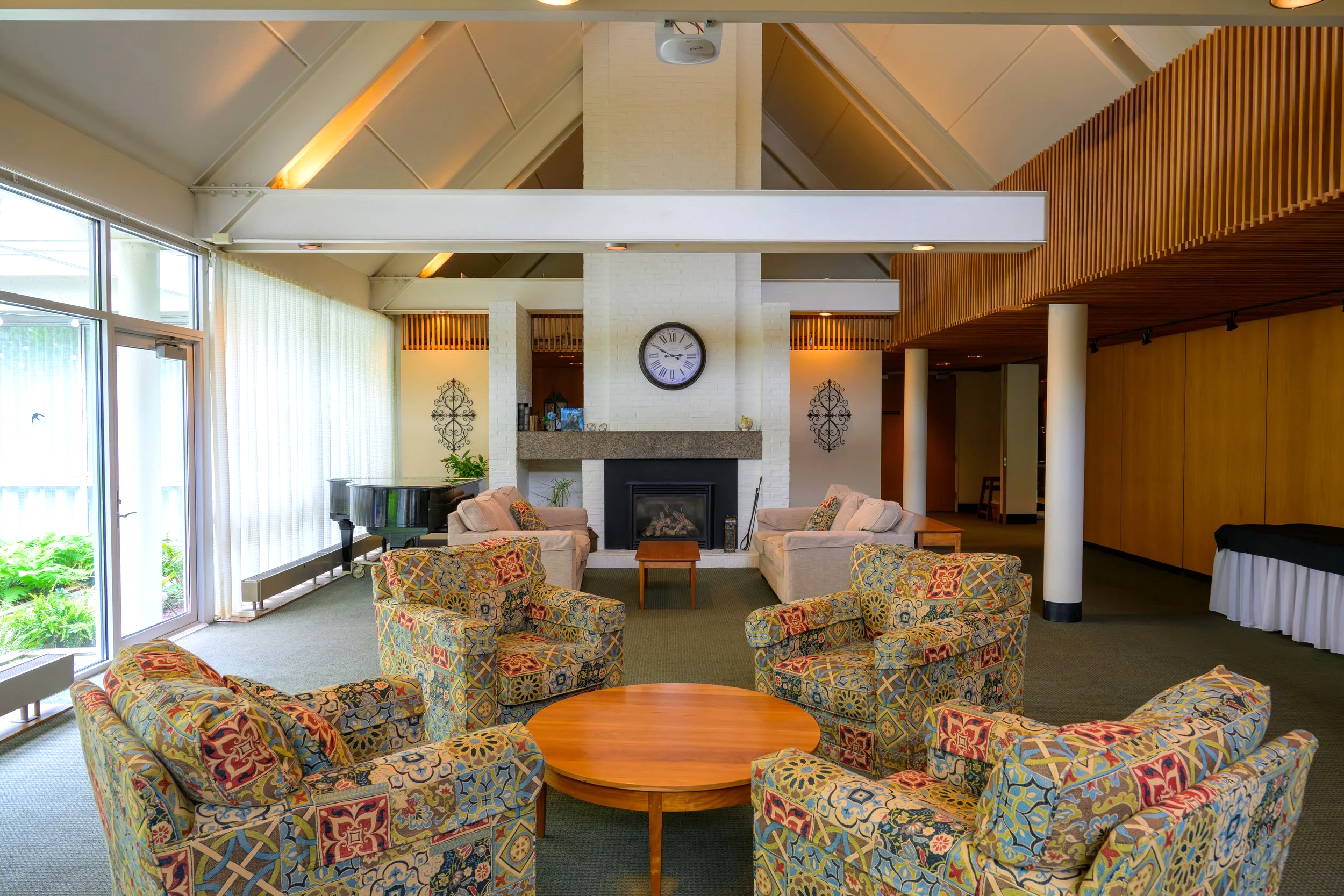 A cozy living room with patterned armchairs arranged in a circle around a wooden coffee table, white brick fireplace with a decorative clock above, large windows with curtains letting in natural light, and modern wood accents on the ceiling and walls