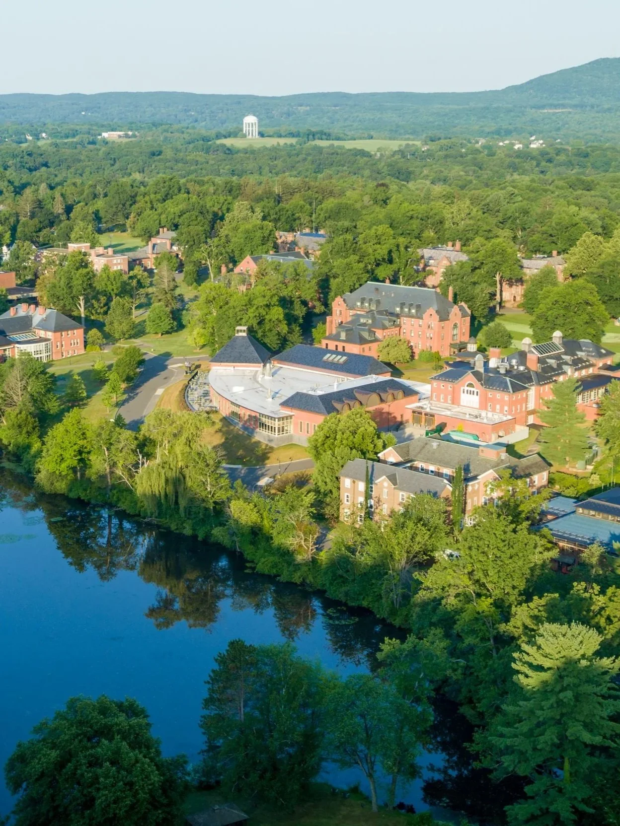 Aerial view of Mount Holyoke College's campus with red brick buildings surrounded by green trees and a pond in the foreground, with hills and a water tower in the distance.
