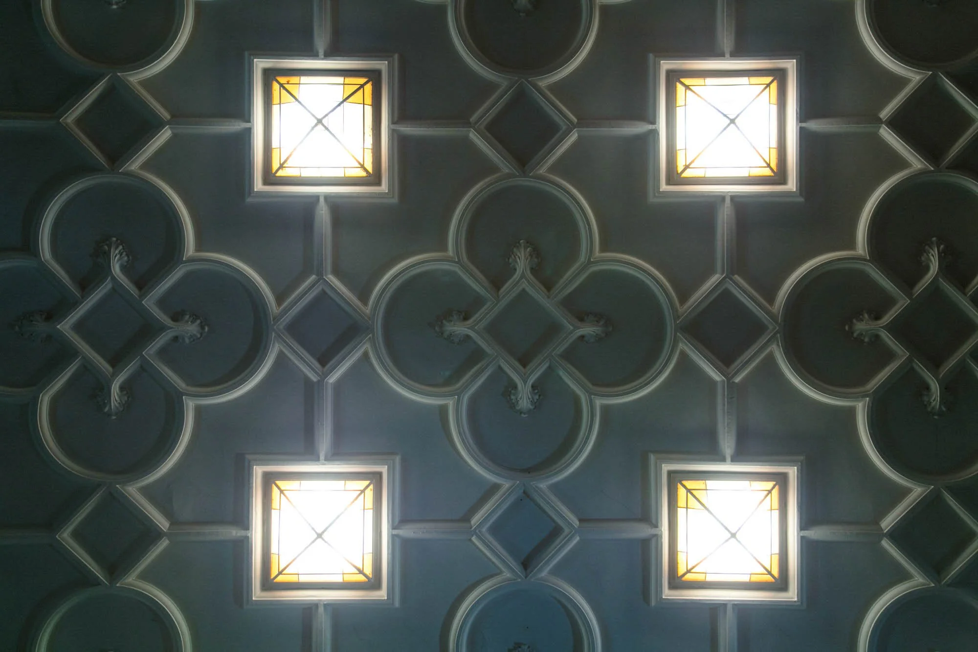 Ceiling with ornate patterns and four square skylights emitting bright light.