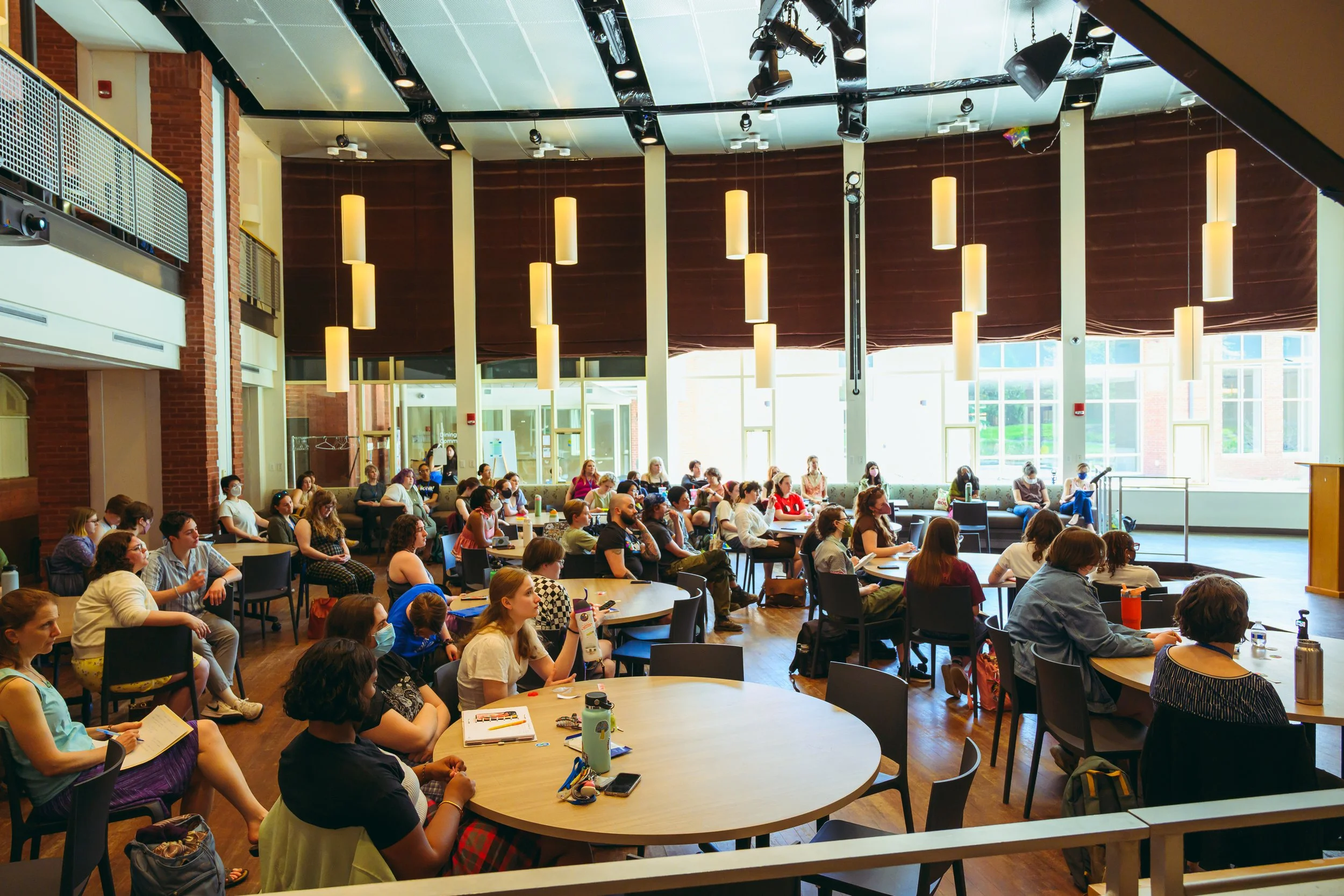A large group of people seated at round tables in a spacious, well-lit conference room, attending a presentation or seminar.