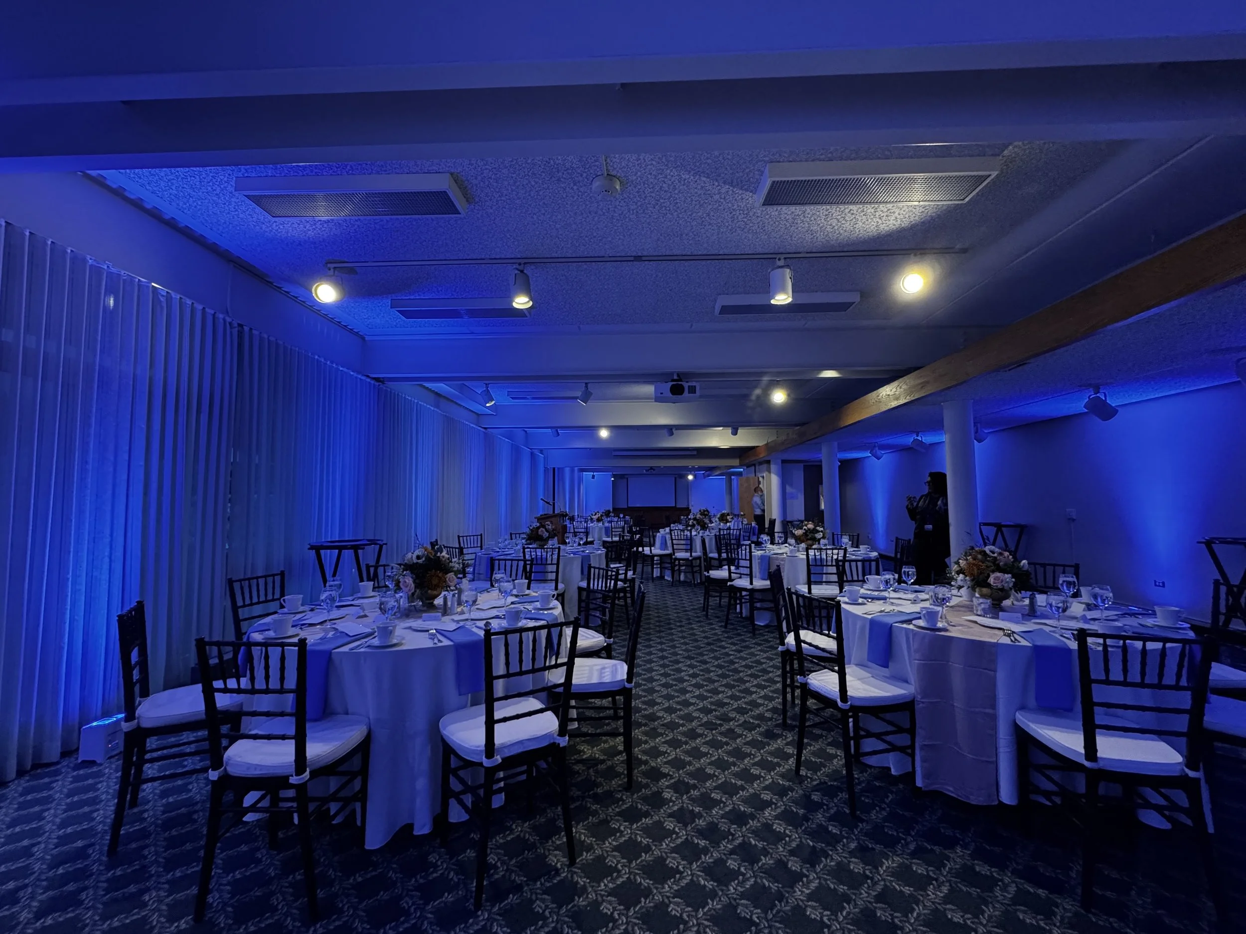 Elegant banquet hall with round tables covered in white tablecloths, decorated with flower arrangements, set for a formal event. The room is illuminated with blue lighting, and there are chairs with white cushions around the tables.