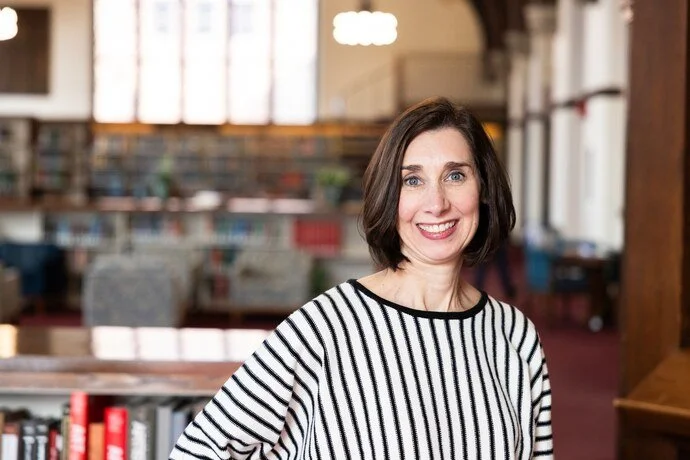 A woman with shoulder-length brown hair and a striped black and white shirt smiling indoors, with bookshelves and large windows in the background.