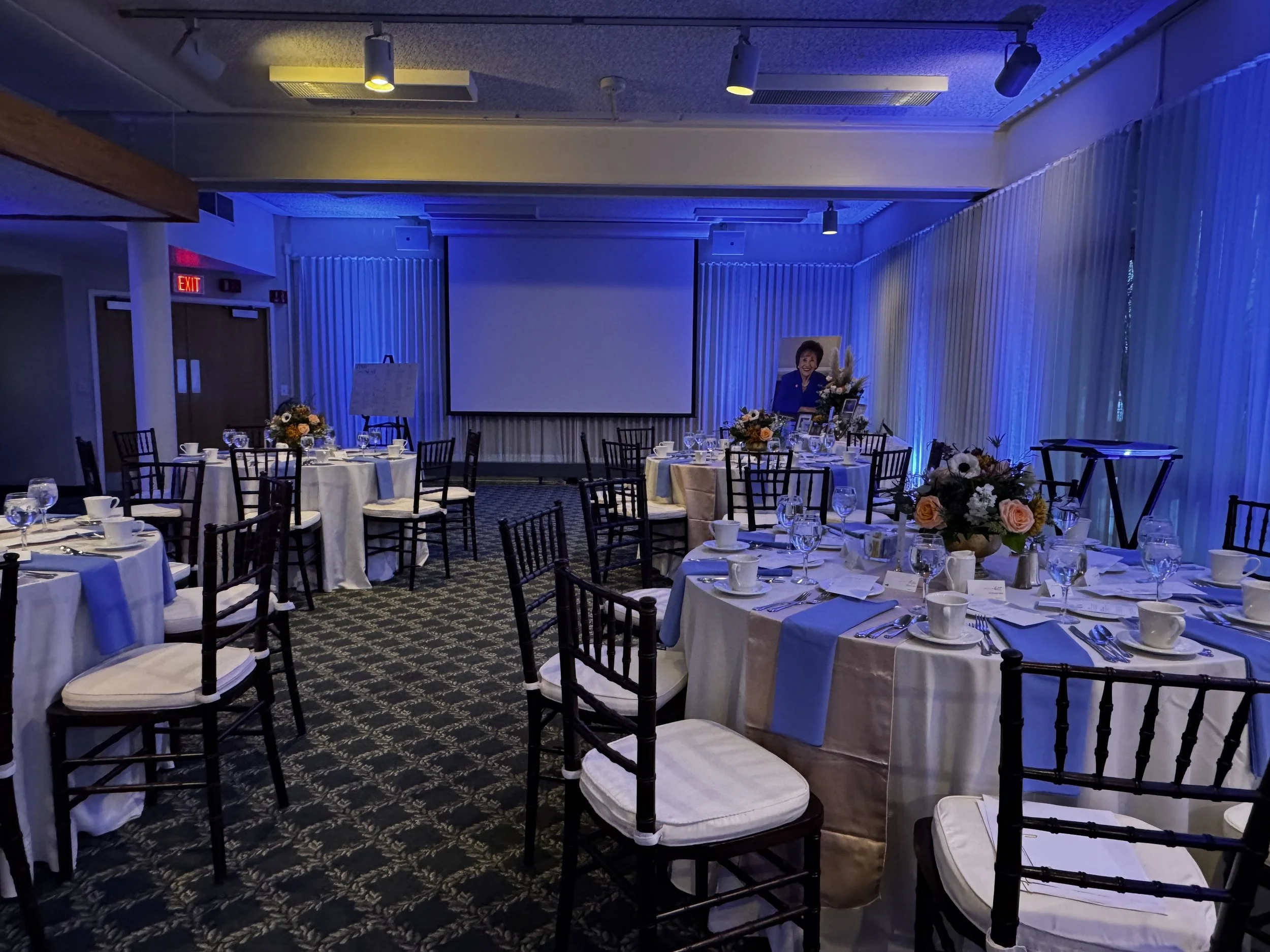 A banquet hall decorated for an event with round tables covered in white tablecloths, floral centerpieces, and place settings, with a large projection screen and a portrait of a woman on display.