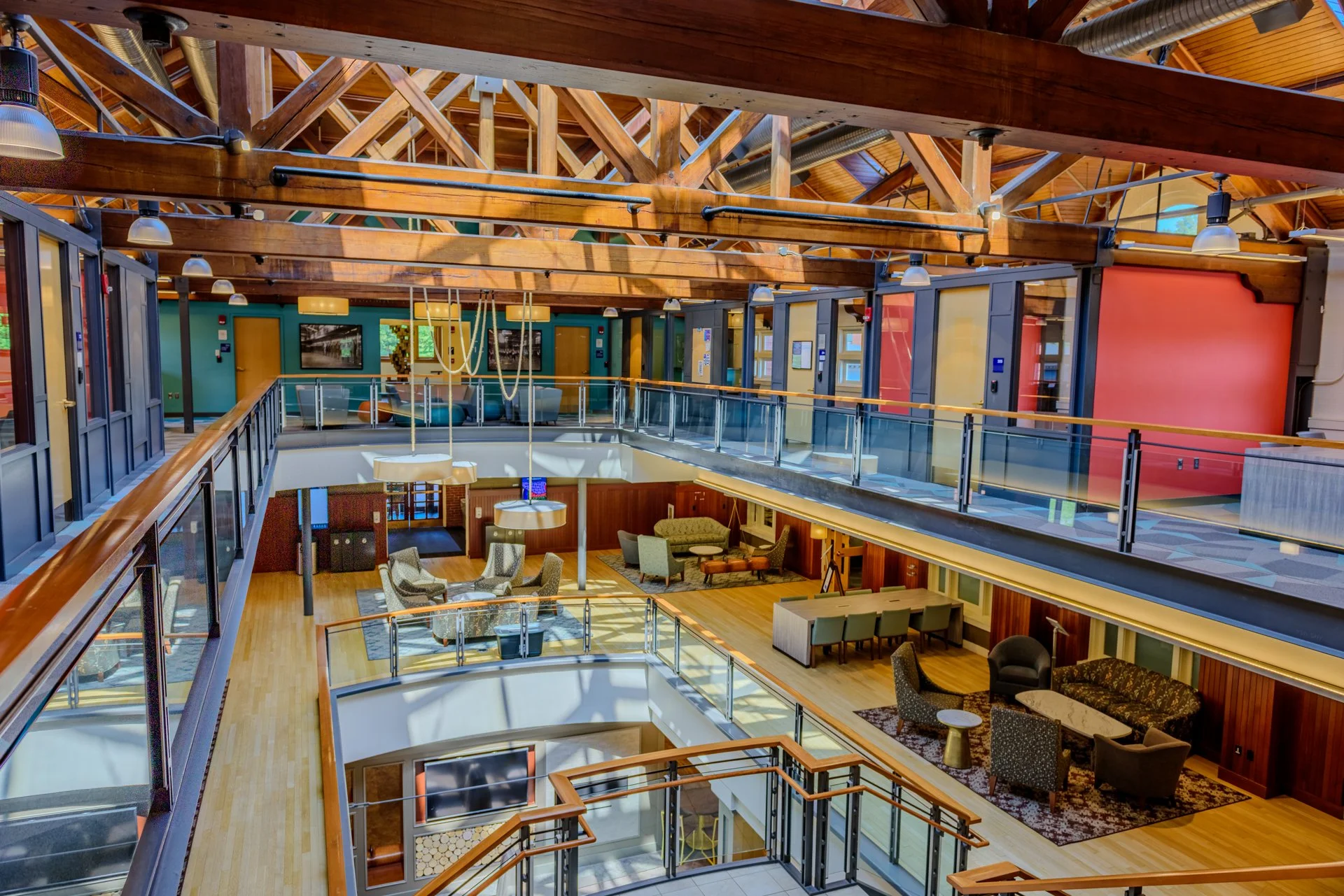 Interior view of a multi-level building with wooden beams, glass railings, and various seating areas including armchairs, sofas, and desks.