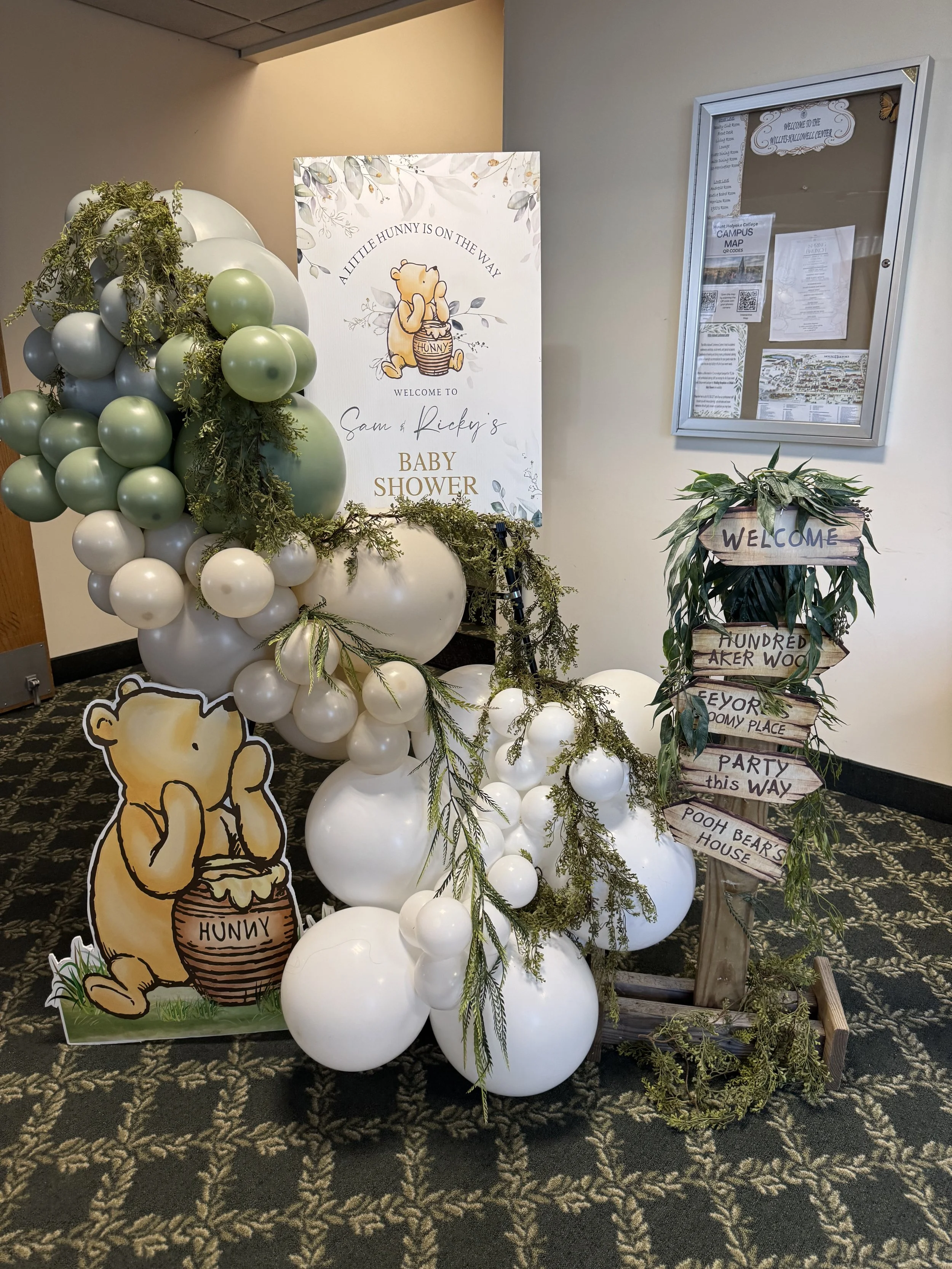 Decorative setup for a baby shower with a balloon arch in shades of white, gray, and green, baby-themed signs, greenery, and a welcome sign for Sam and Ricky's baby shower. There is a cartoon illustration of Winnie the Pooh holding a honey jar with t