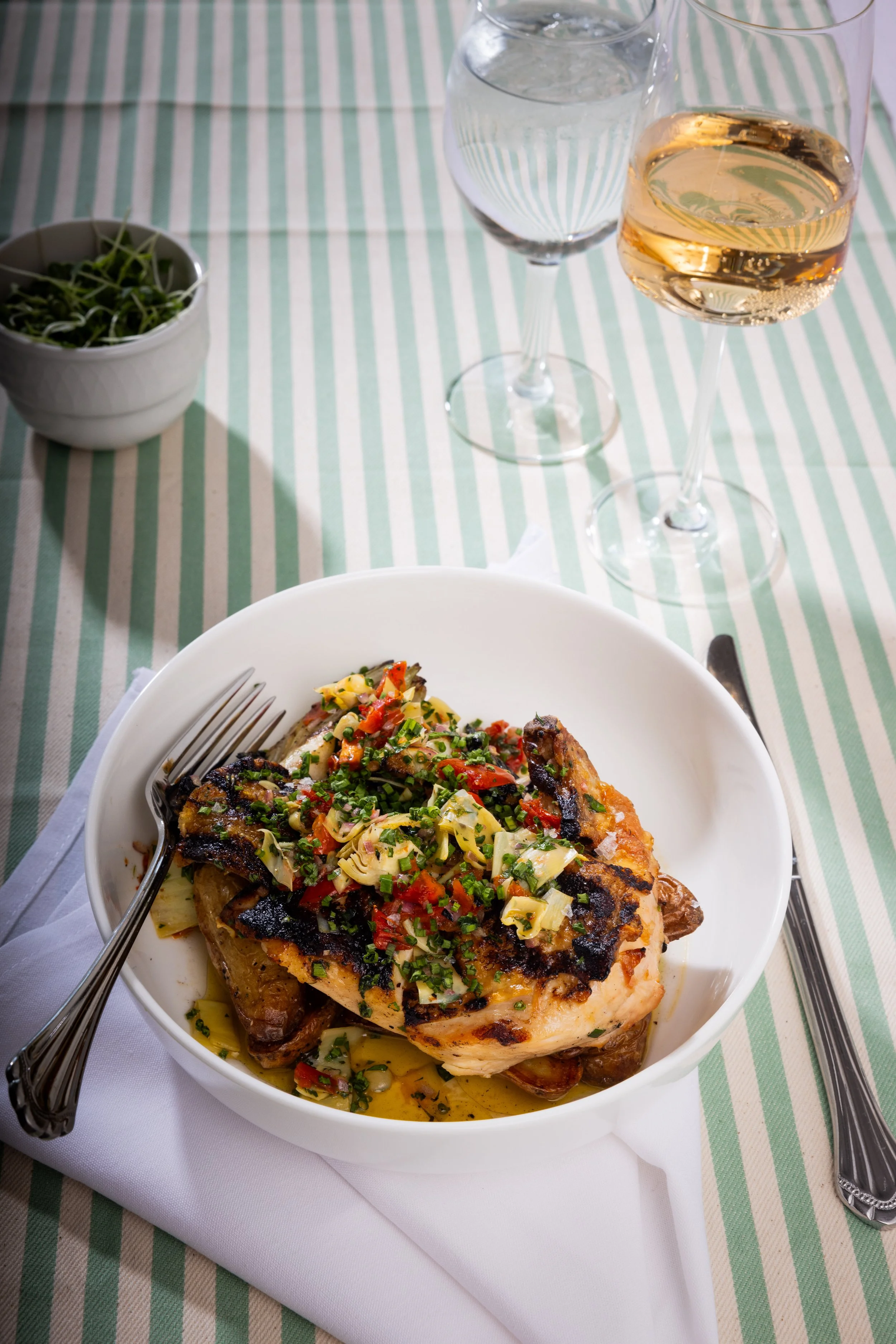 A white bowl filled with grilled chicken topped with colorful chopped vegetables and herbs, sitting on a white plate with a fork and knife. In the background, there are two glasses of water and one glass of rosé wine on a green and white striped tabl