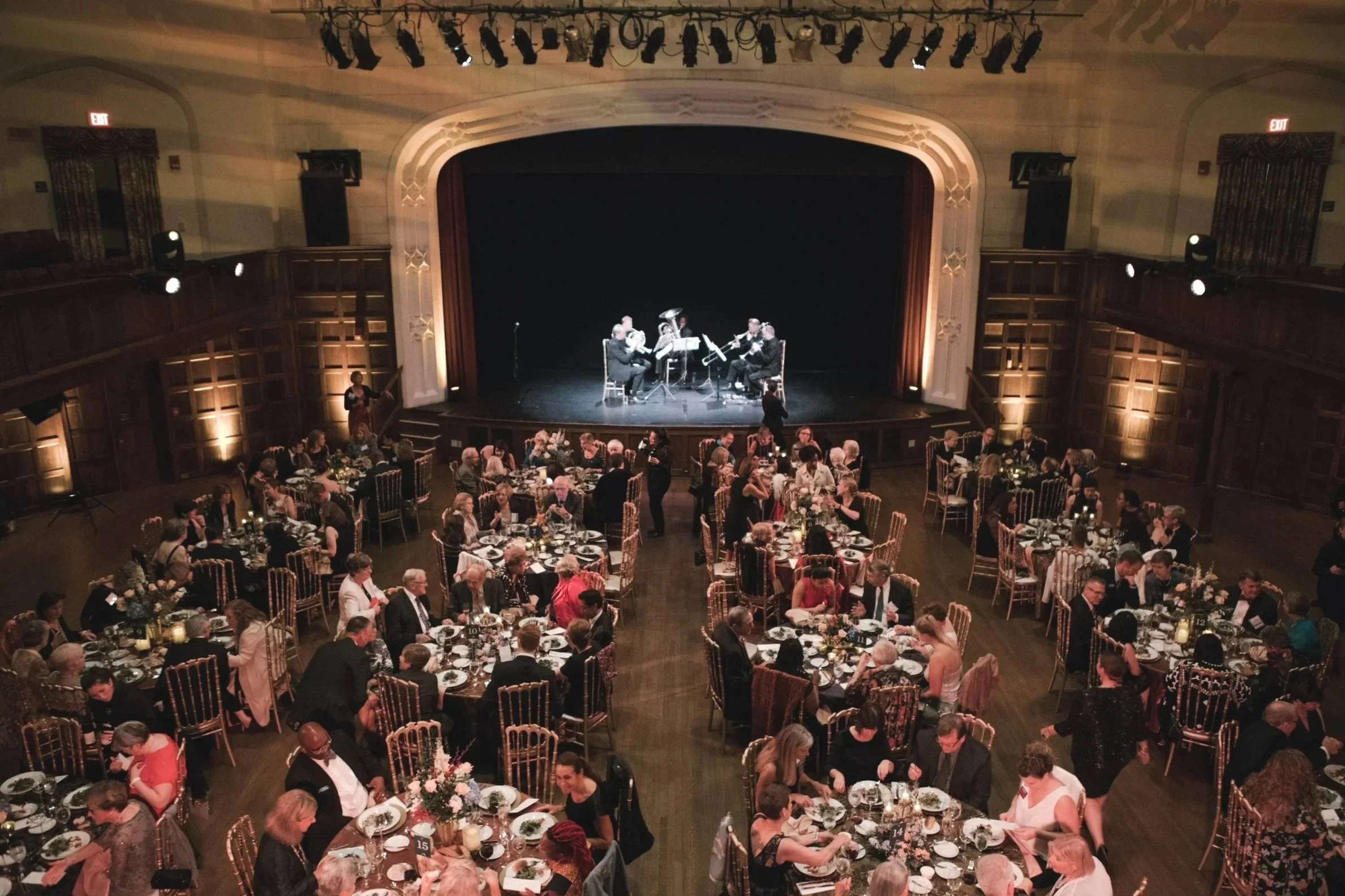 A large banquet hall filled with many round tables set for a formal dinner event, with numerous people seated and dining. On a stage at the front, four musicians are performing, surrounded by a dark background. The hall features ornate woodwork and s
