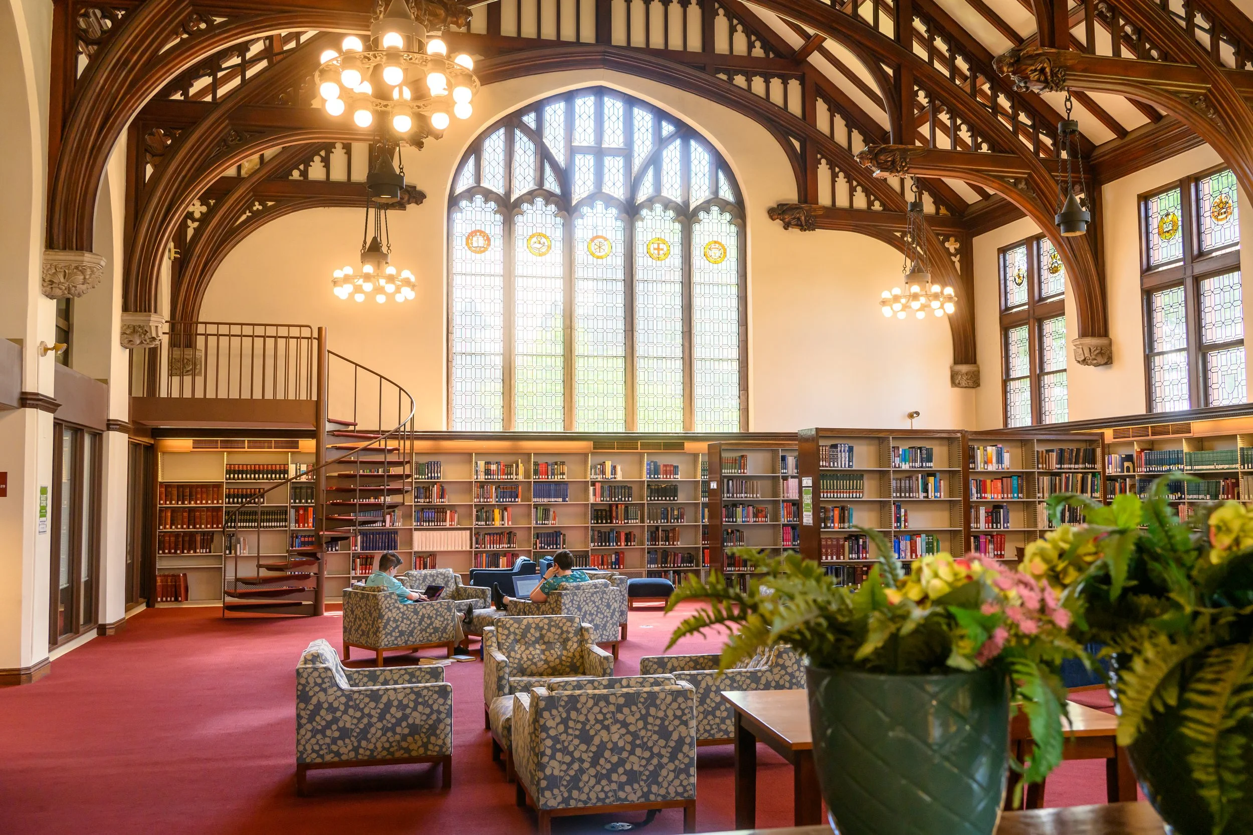 Interior of a historic library with high vaulted windows, wooden arches, and chandeliers. There are bookshelves, armchairs, and two children reading.