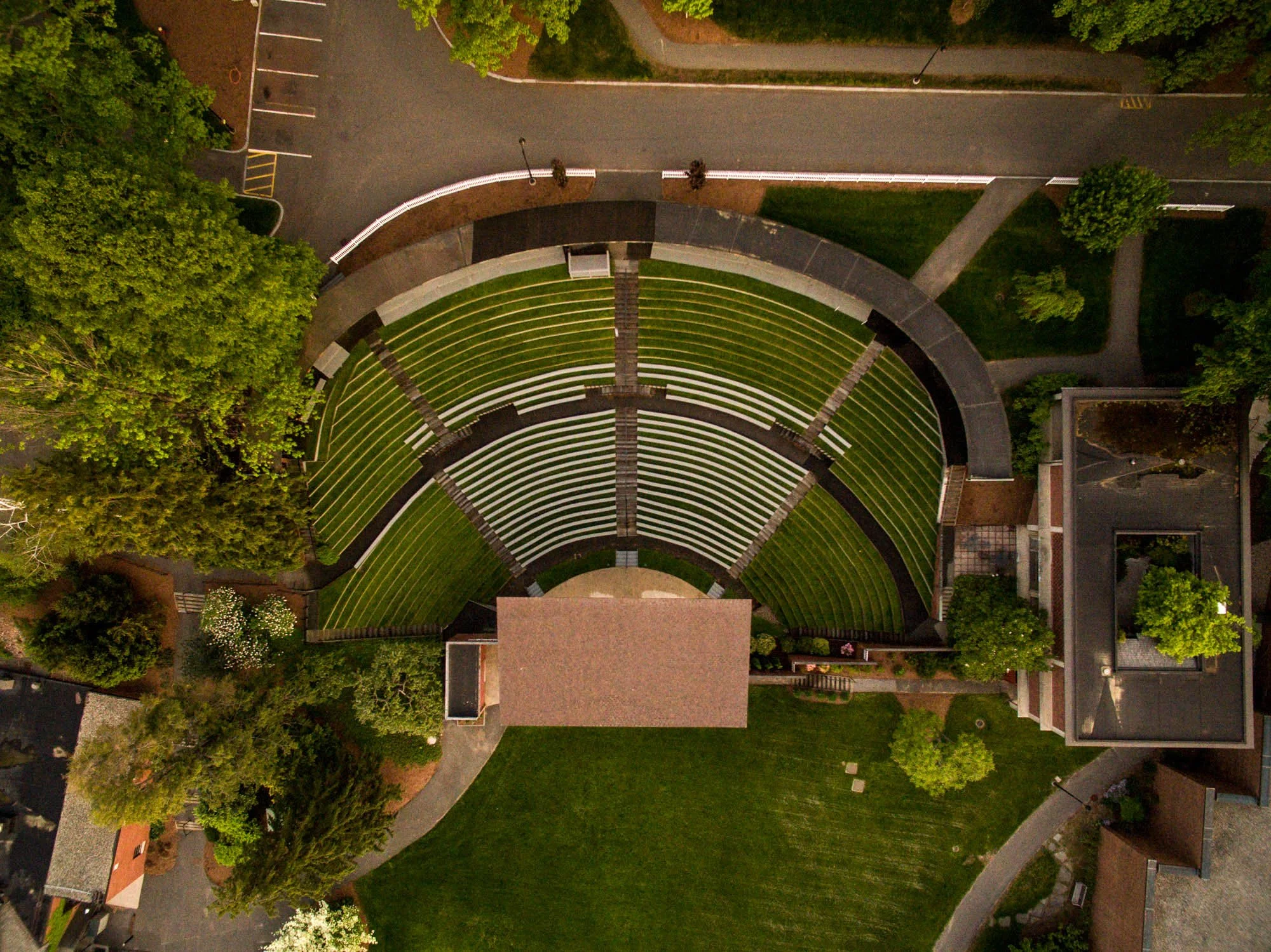 An aerial view of an outdoor amphitheater with a curved seating area, surrounded by trees and nearby residential buildings.