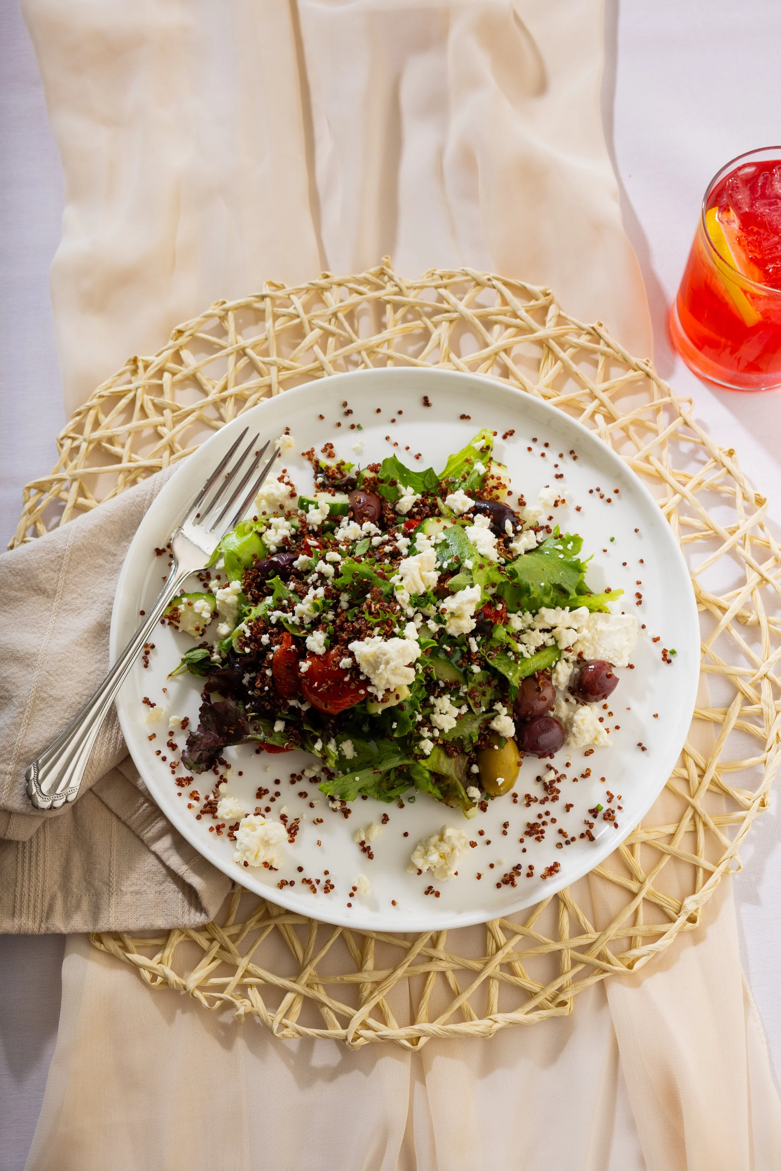 A plate of salad with greens, cherry tomatoes, olives, feta cheese, and red quinoa, with a fork resting on a napkin, on a woven placemat, and a glass of pink beverage with lemon slices.
