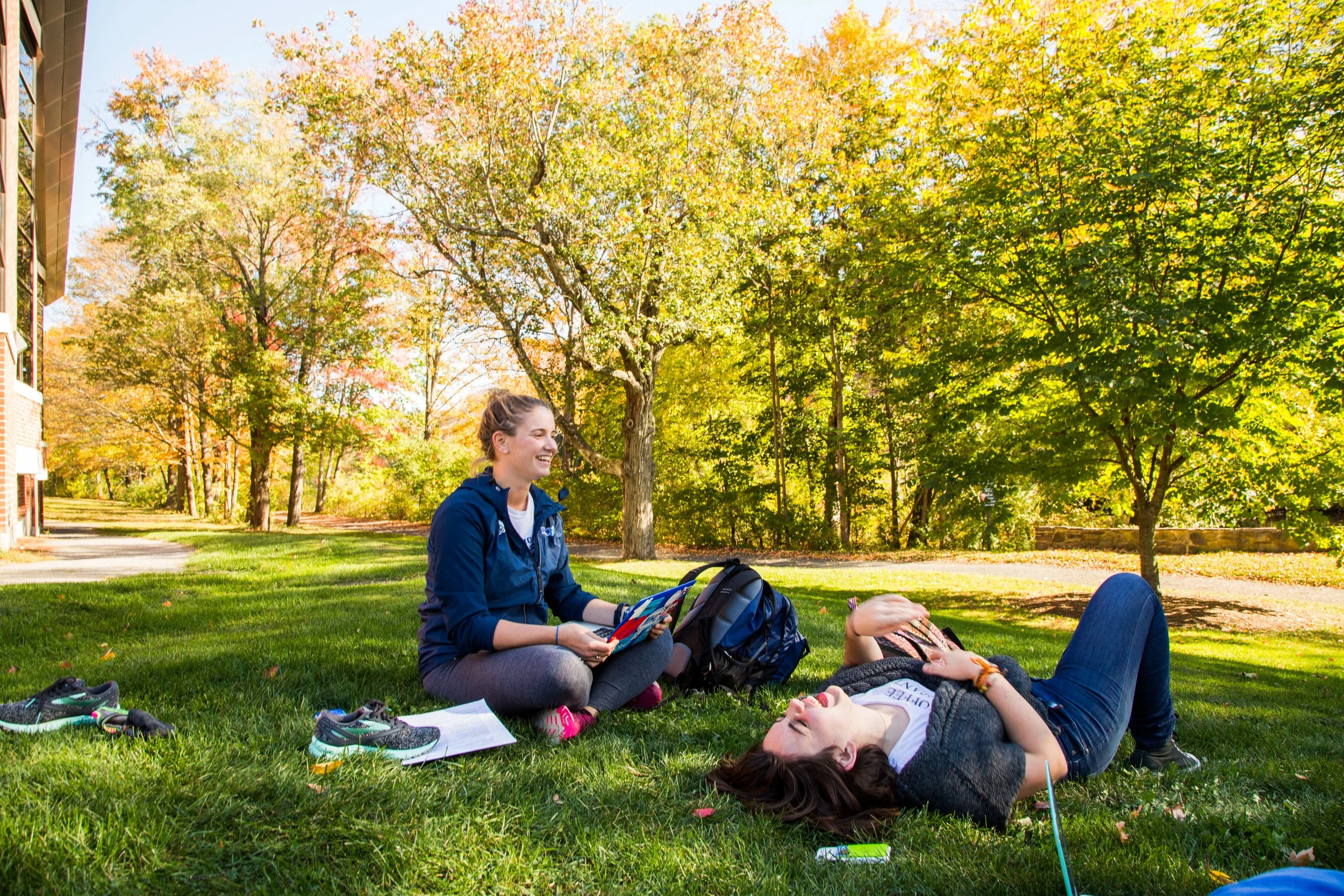 Two young women relaxing on the grass in a park during autumn, with trees and a building in the background. One woman is sitting, holding a book, and smiling, while the other is lying down with sunglasses, laughing, and covering her face with her hand.