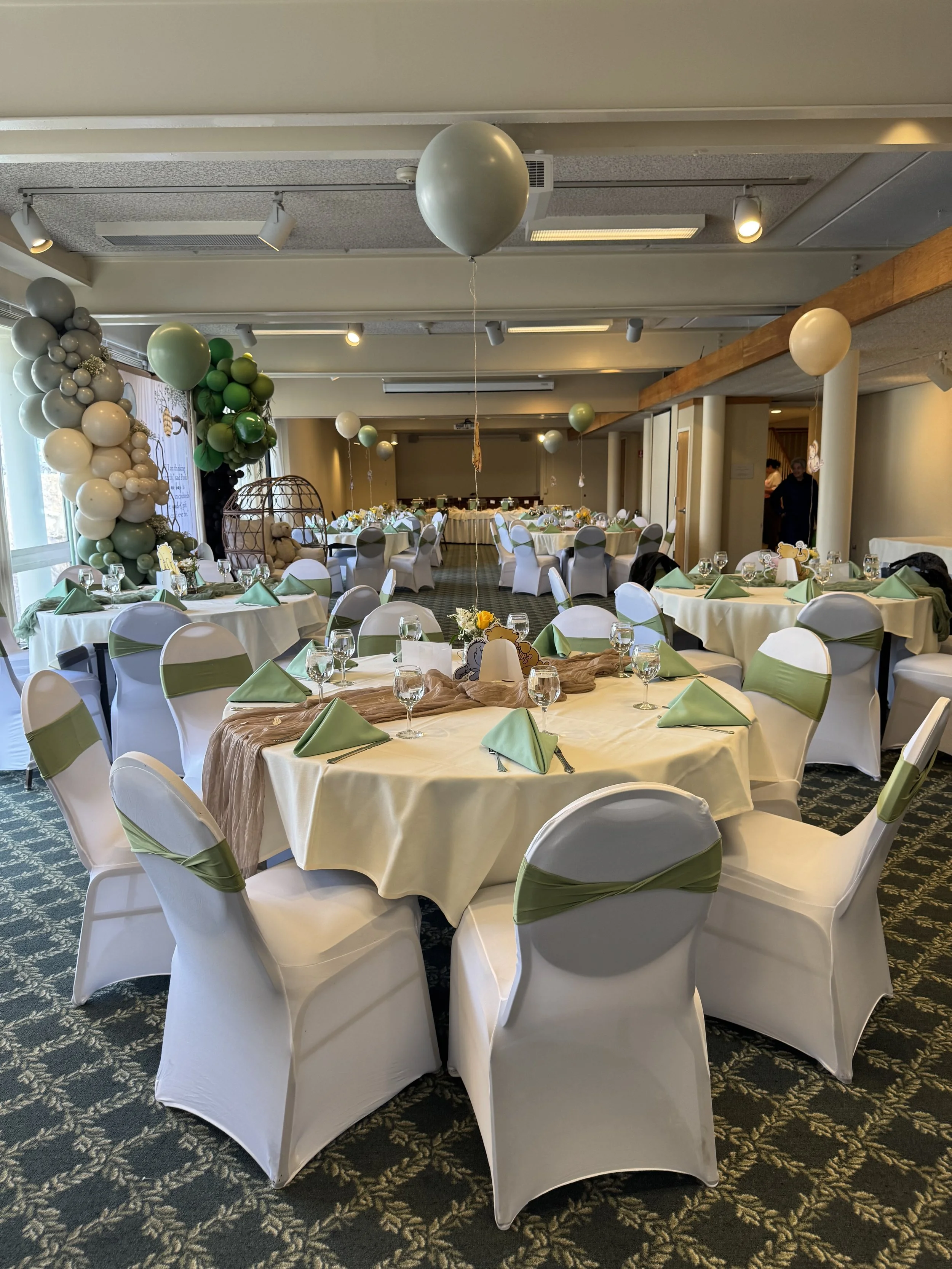 A decorated banquet hall with round tables covered in white tablecloths and green napkins, surrounded by white chairs with green sashes. Balloons and a balloon arch decorate the room, with a sitting area and a few people visible in the background.