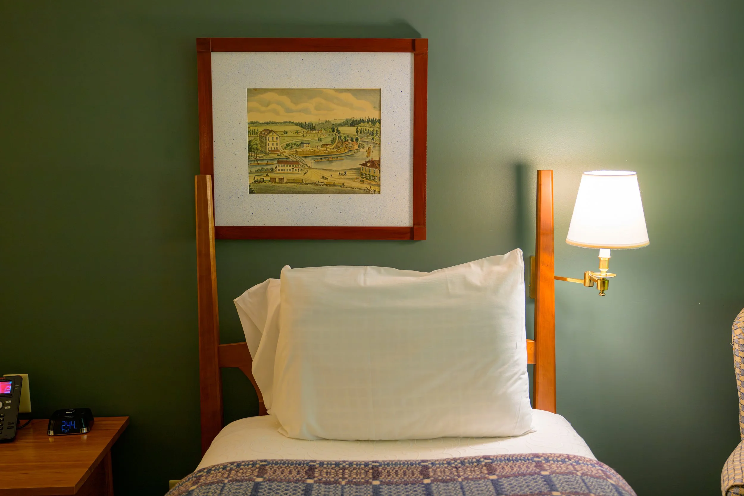 Hotel room with a bed, resting against a green wall, with a wooden headboard, a pillow, a framed painting of a landscape, a bedside lamp, and a nightstand with a clock and a phone.