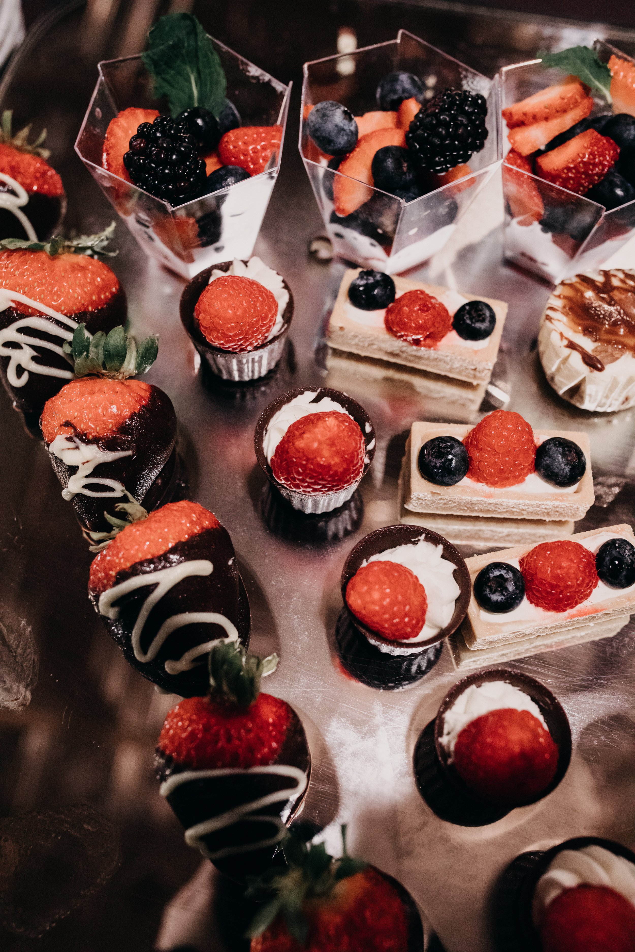 Assorted desserts topped with fresh berries, including strawberries, blueberries, blackberries, and raspberries, arranged on a metallic tray.
