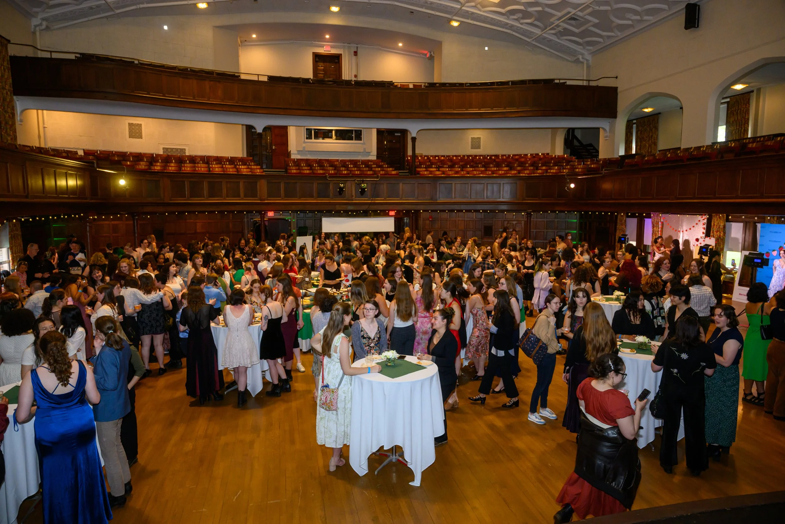 Crowd of people attending a social event or gathering in a large room with wooden floors, tables, and seating. The room has a balcony area and decorative curtains, with some people standing and chatting while others are seated.