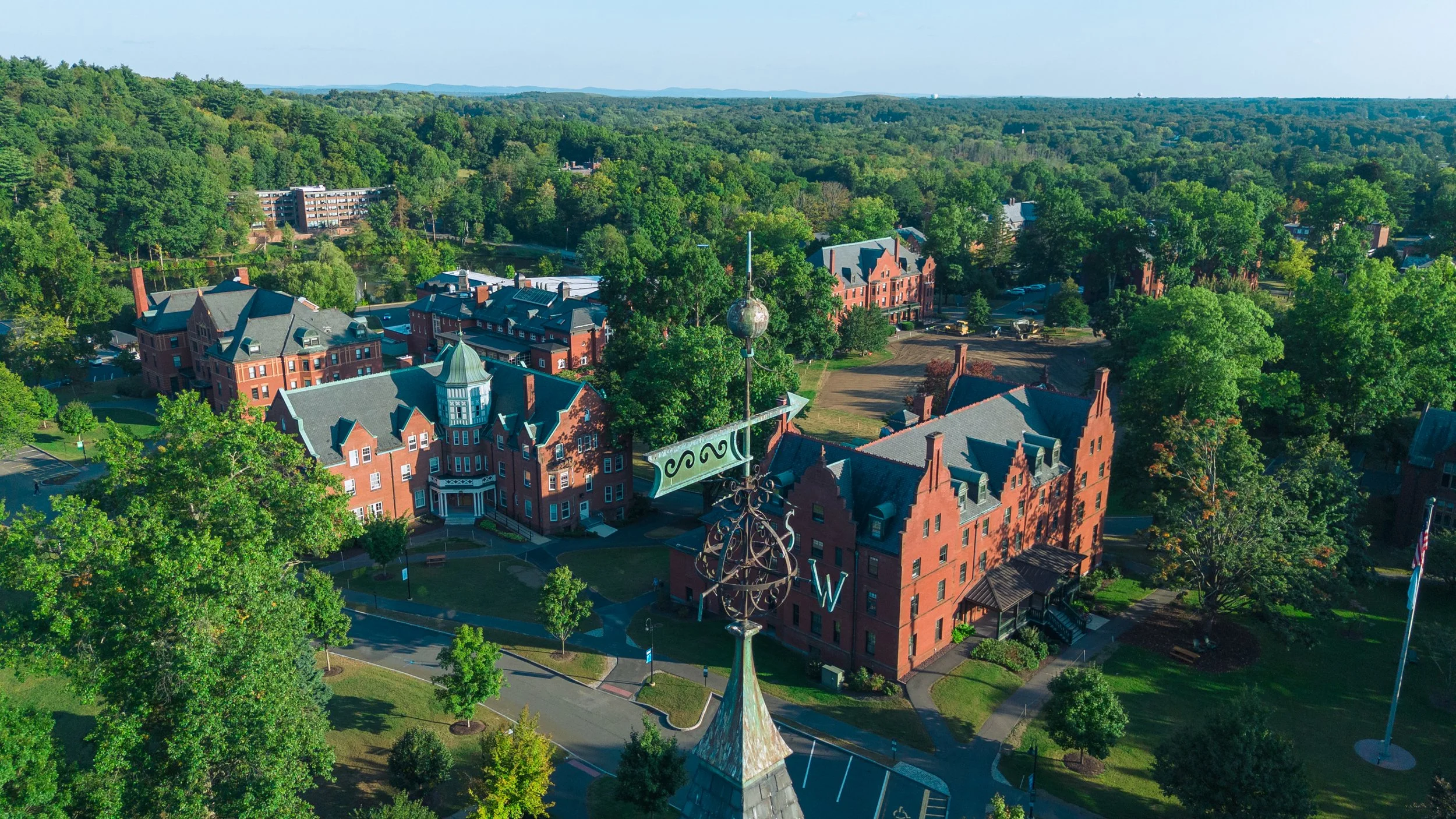 Aerial view of a historic campus with red brick buildings, surrounded by green trees and rolling hills in the distance.