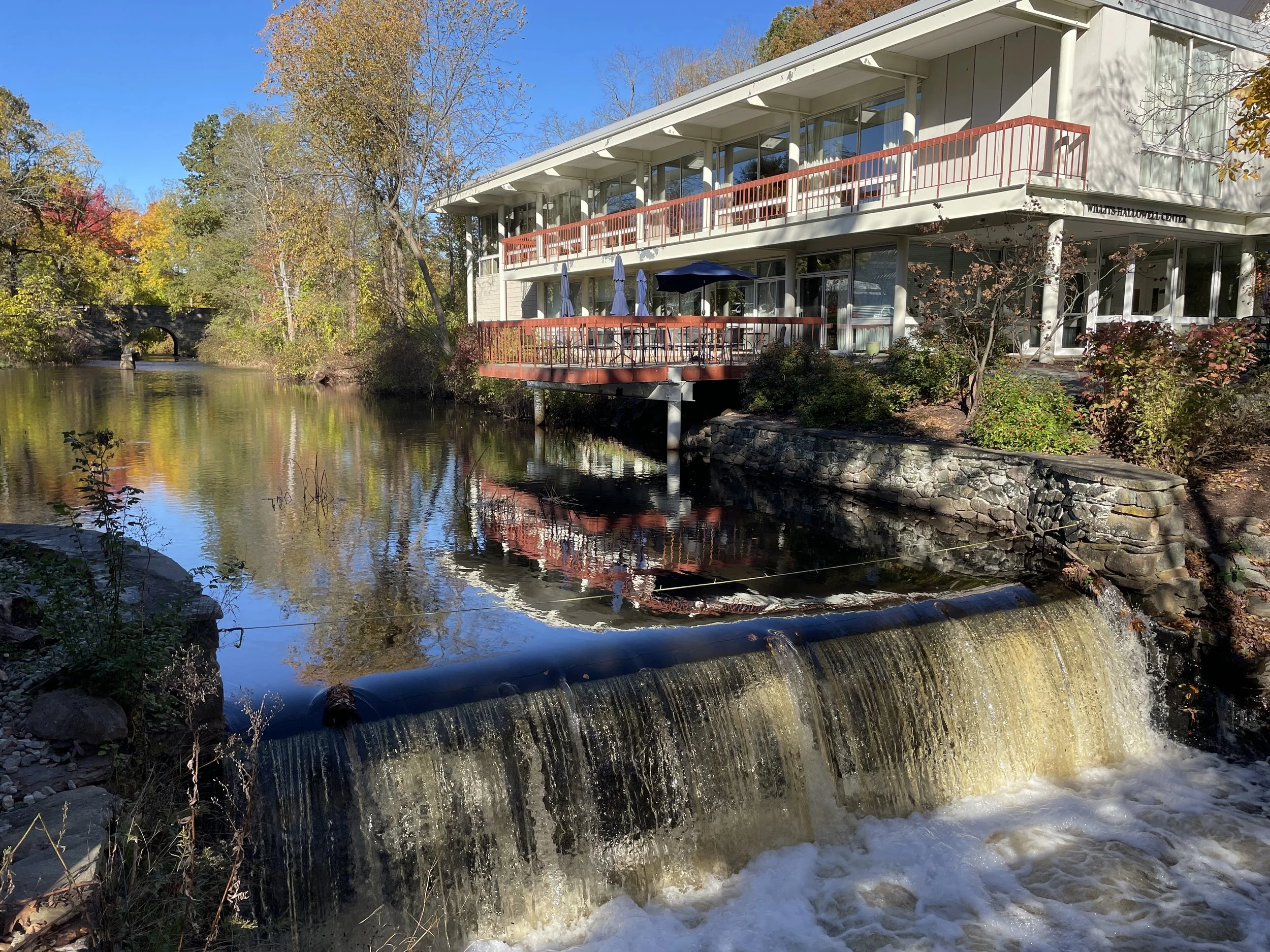 A modern multi-level building with glass windows and balconies by a river, surrounded by colorful autumn trees, with a stone bridge visible in the background and a small waterfall in the foreground.