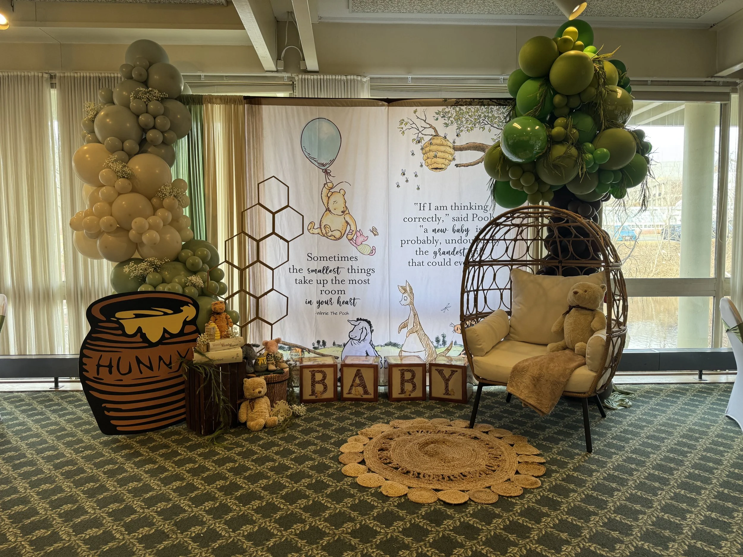 Baby shower decorations with green and beige balloons, Winnie the Pooh theme, a plush teddy bear on a sofa, a woven rug, and a backdrop with Winnie the Pooh and related quotes.