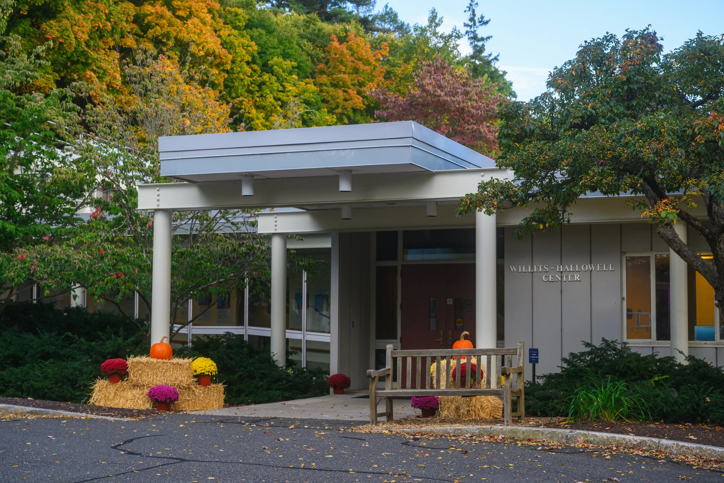 The entrance of the Willits-Hallowell Center decorated for fall with hay bales, pumpkins, and colorful chrysanthemums, surrounded by trees with autumn foliage.
