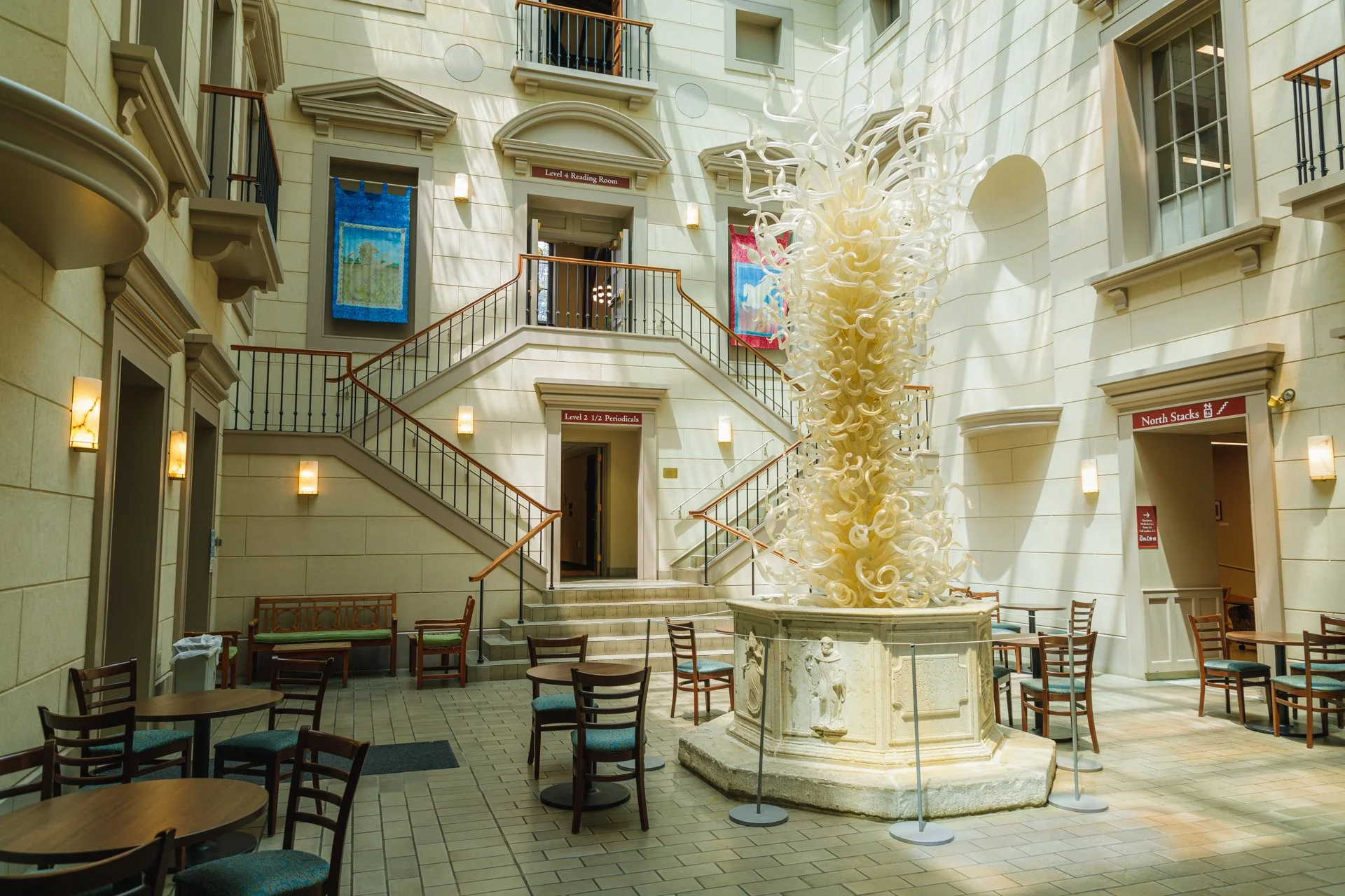 Interior view of a hotel lobby with a large decorative glass sculpture in the center, beige walls, multiple balconies, and chairs and tables arranged for seating.